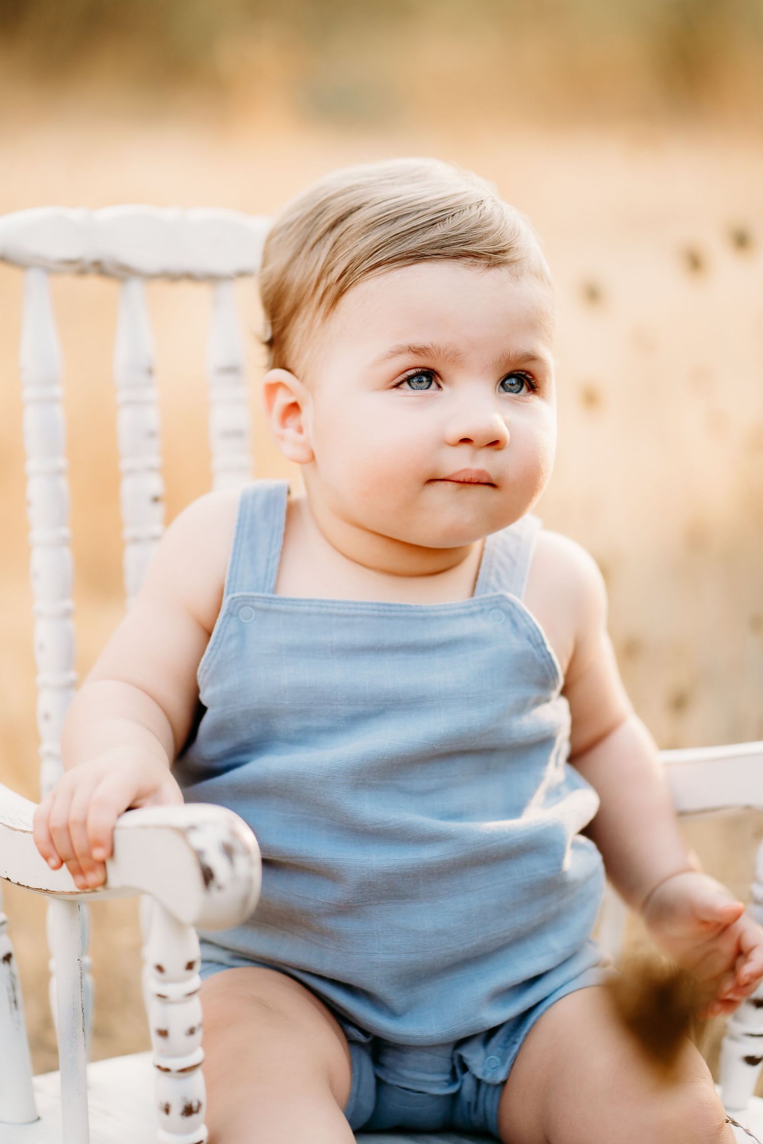 A baby boy wearing a light blue sleeveless romper sits in a white wooden rocking chair in golden field for 9 month photo session.