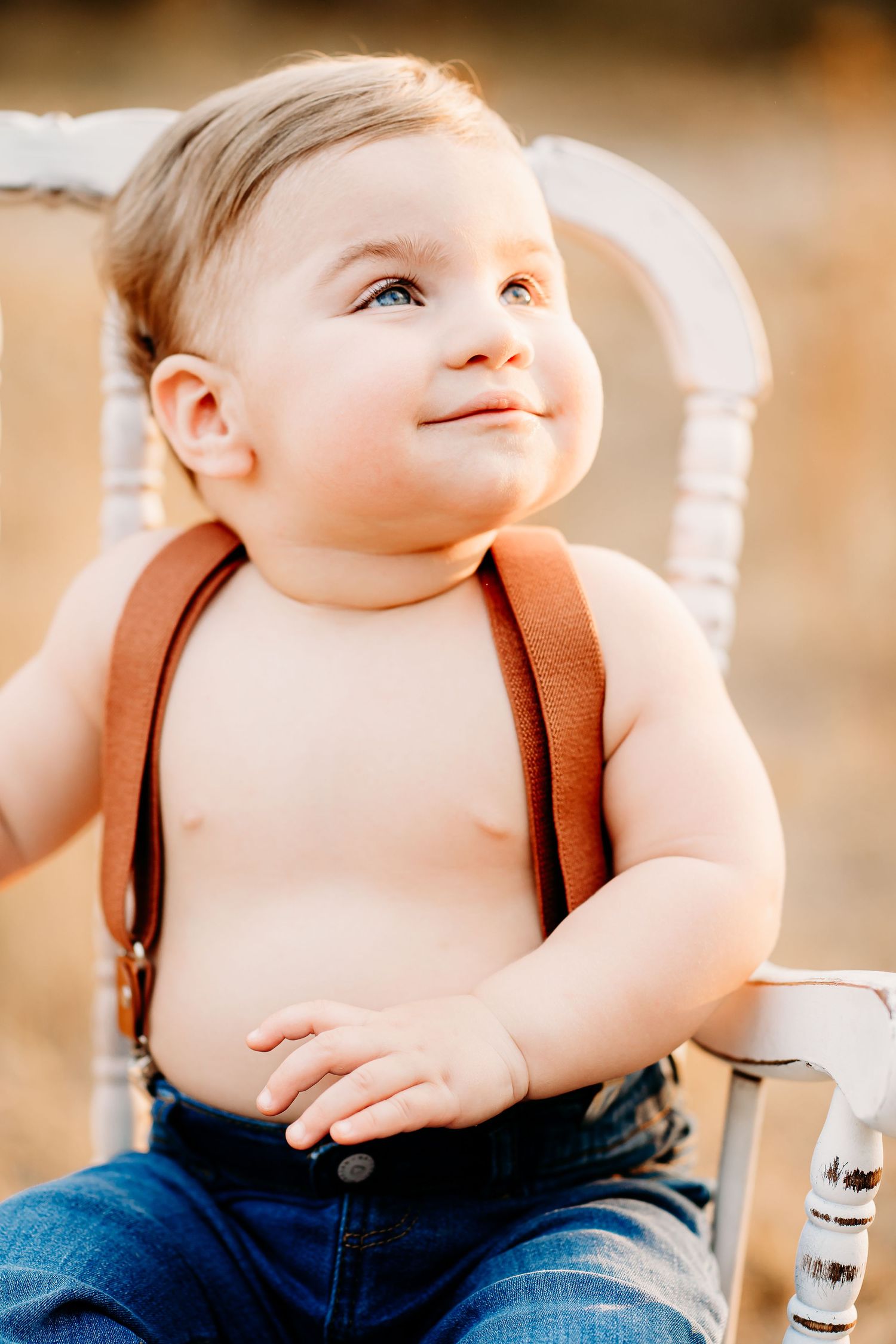 A baby wearing suspenders and jeans sits looking upward while smiling warmly against a blurred background at first birthday photoshoot.