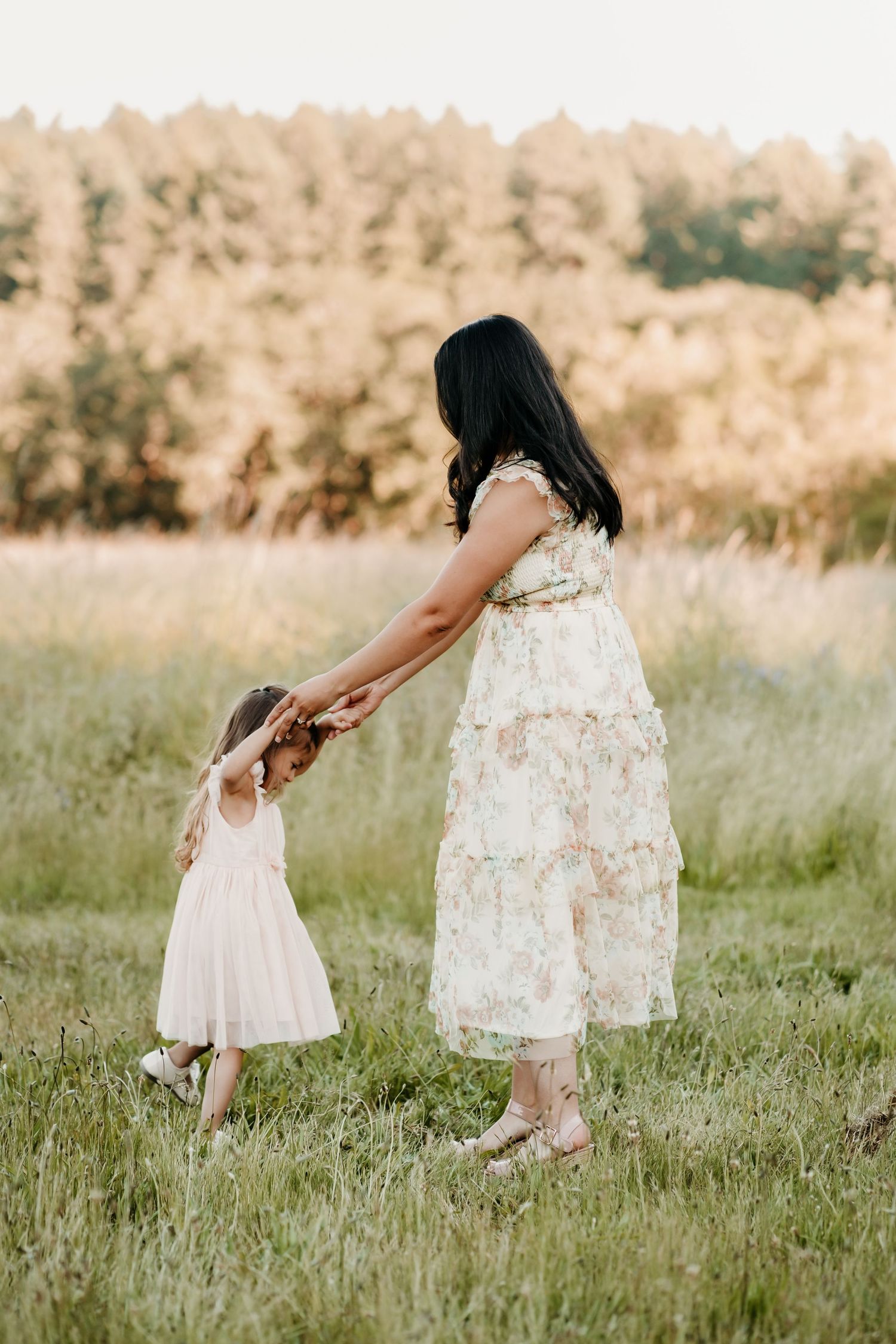 A mother in a floral dress holds her young daughter's hand in a sunlit green field with trees in the background during family photos.