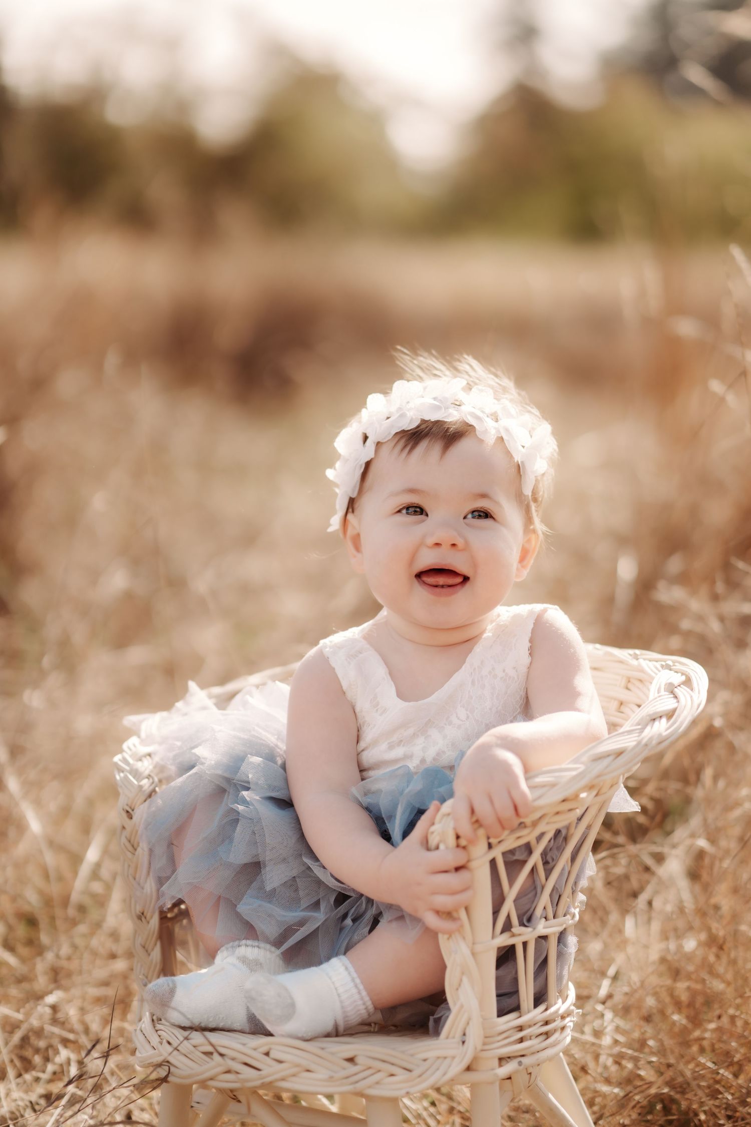 Smiling baby girl in white lace top and blue tutu skirt sitting in wicker chair outdoors in golden field during first birthday photos.