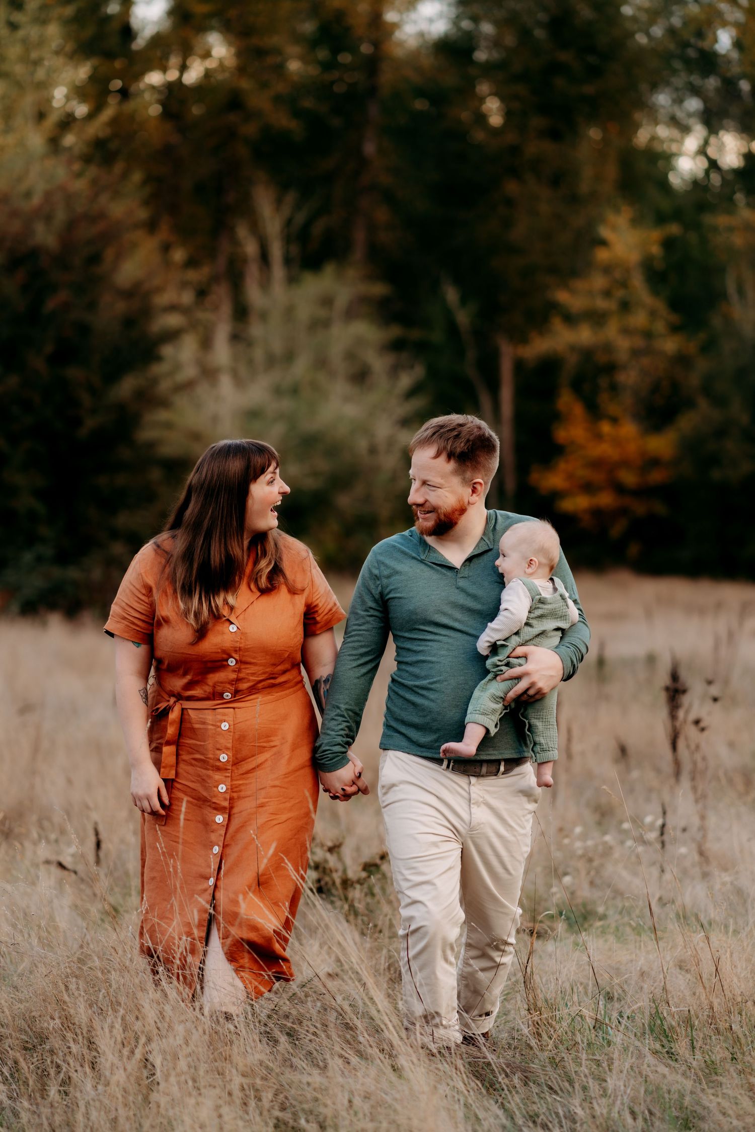 A happy family walks through an autumn field, mother in orange dress and father holding baby in green outfit.