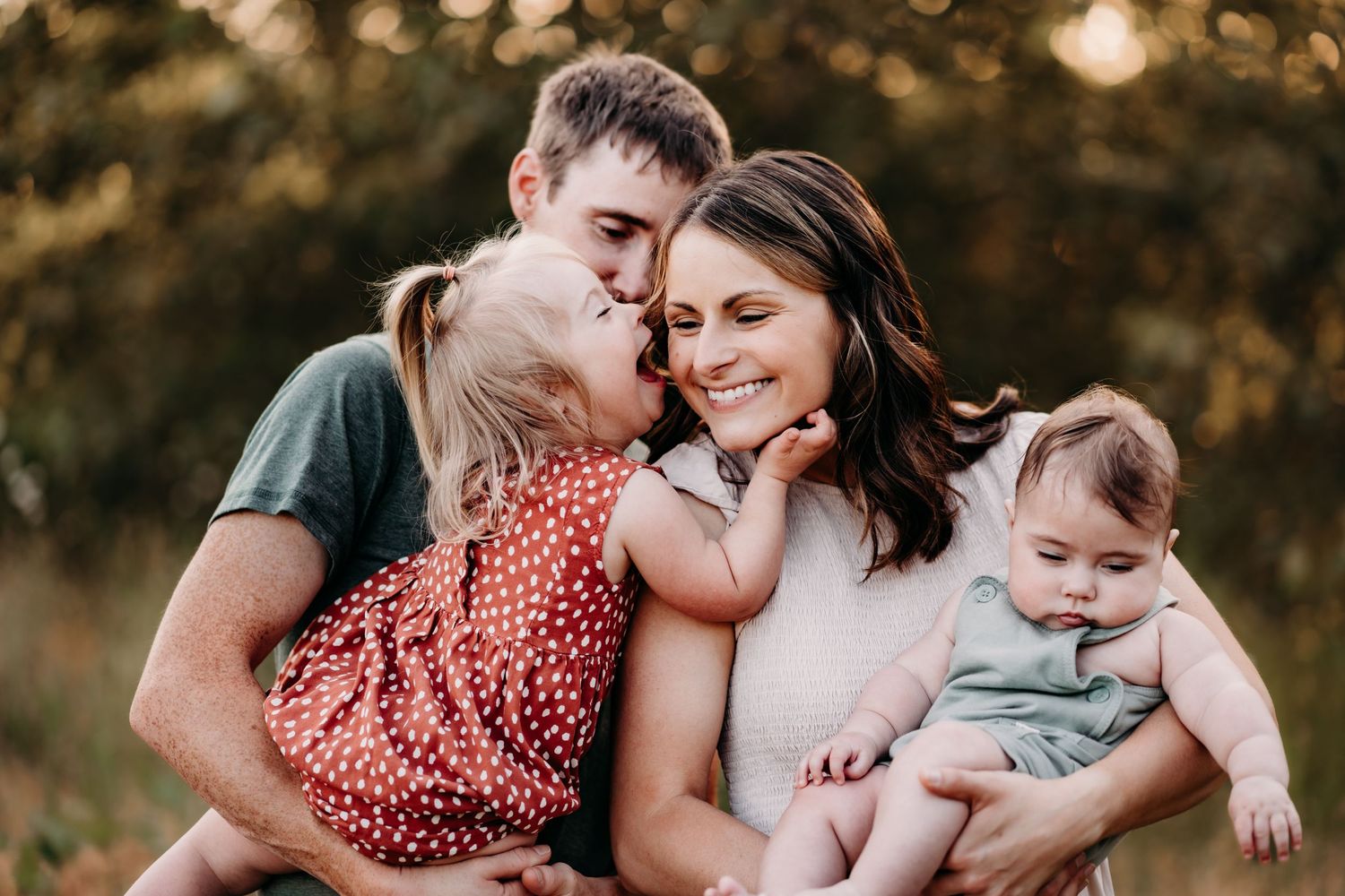 Happy family portrait with parents and two young children sharing kisses outdoors in golden light during family photos in Puyallup, Wa.