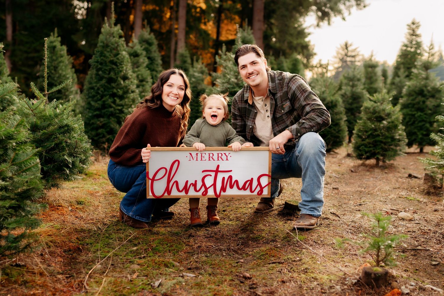 Family of three posing with a Merry Christmas sign at a Christmas tree farm for Christmas photo session in Puyallup, Wa.