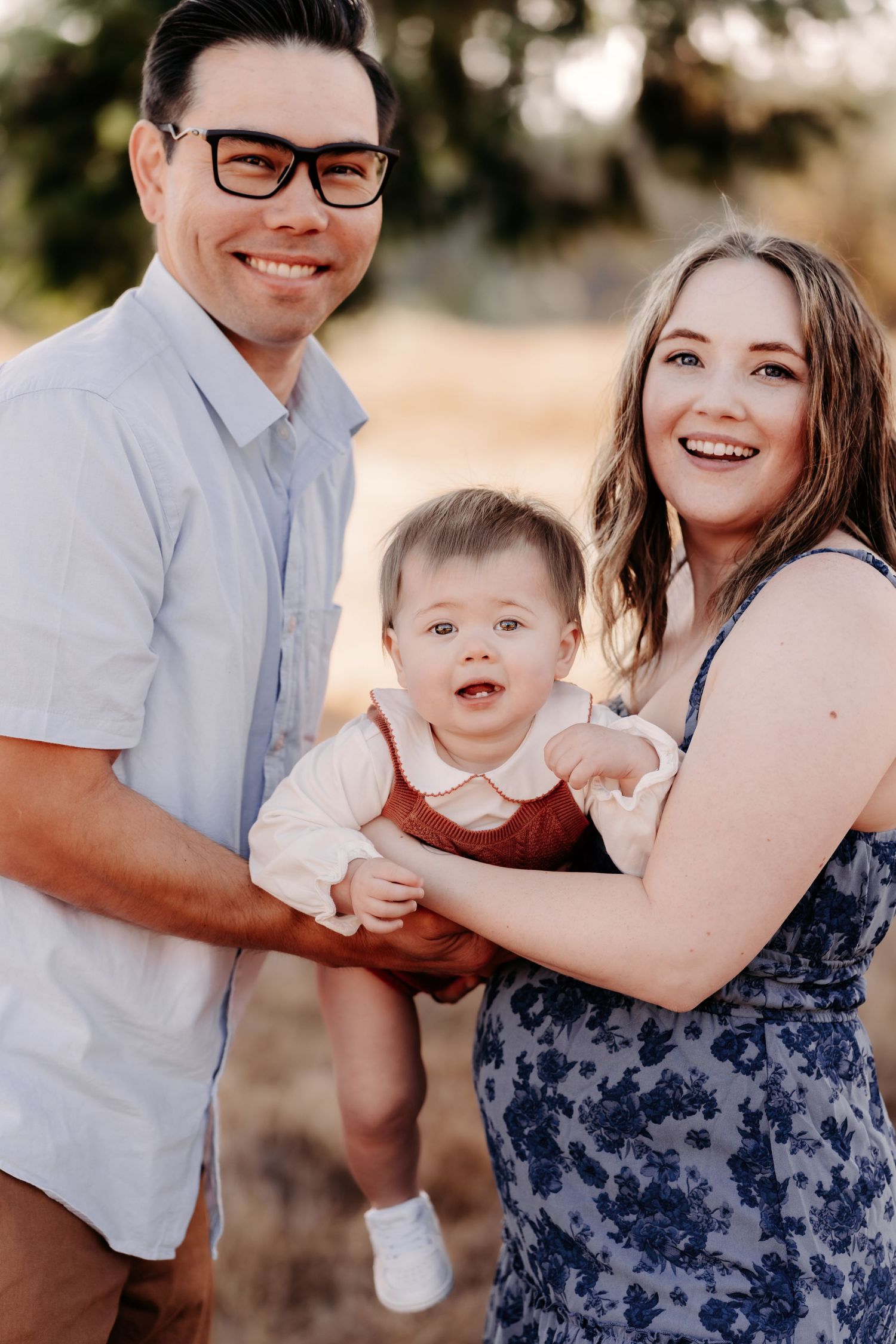 Happy young family with man in glasses, woman in floral dress, and baby in red overalls posing outdoors for family photos in Puyallup, WA