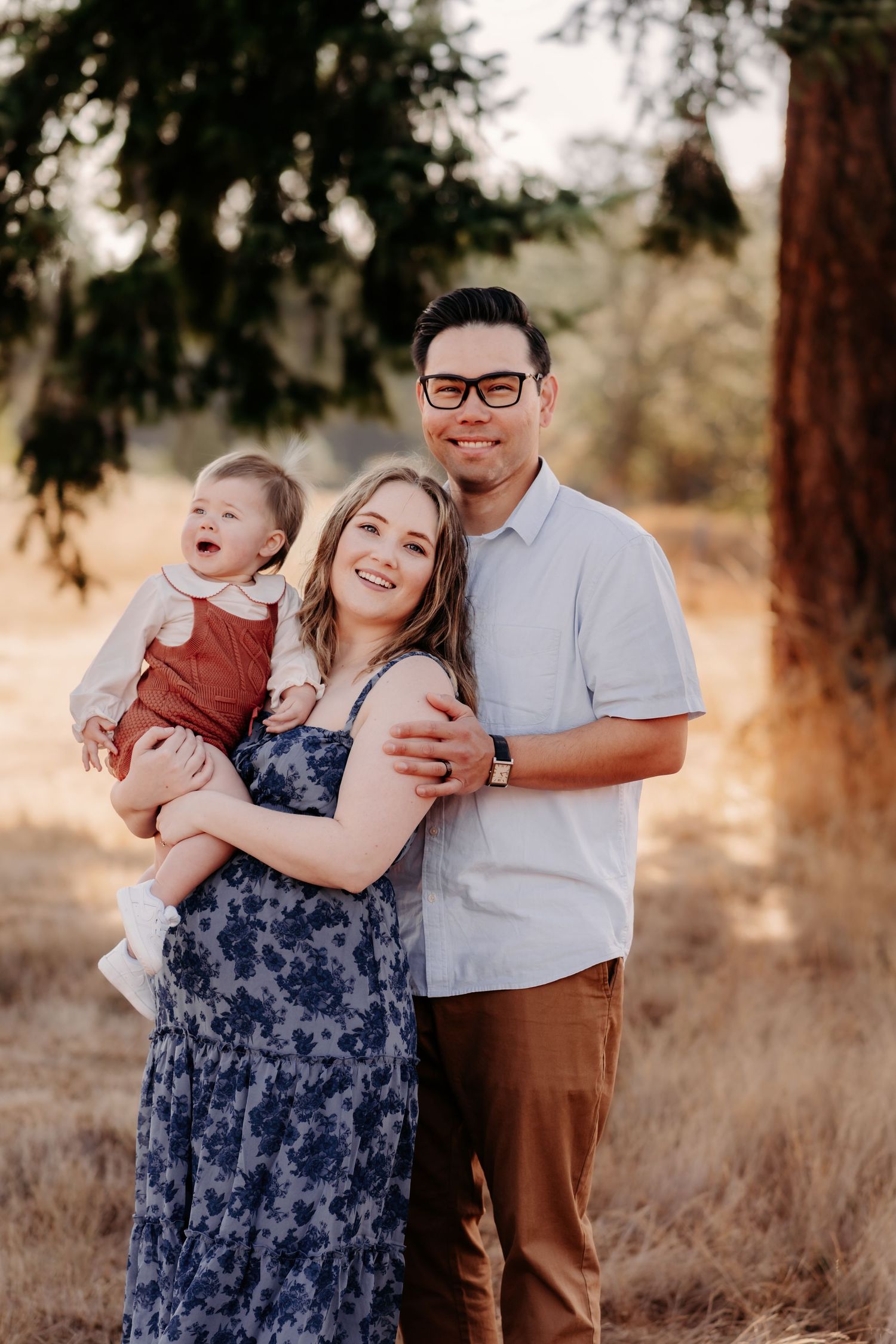 Happy family portrait outdoors, parents smiling and holding a baby in a sunlit field with trees for family photos in Tacoma, WA.