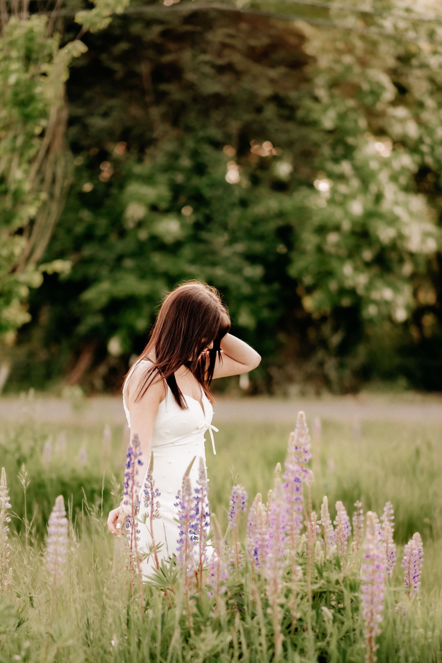 Young woman in white dress standing among purple lupine flowers in a lush green garden field for senior photos in Tacoma, Wa.