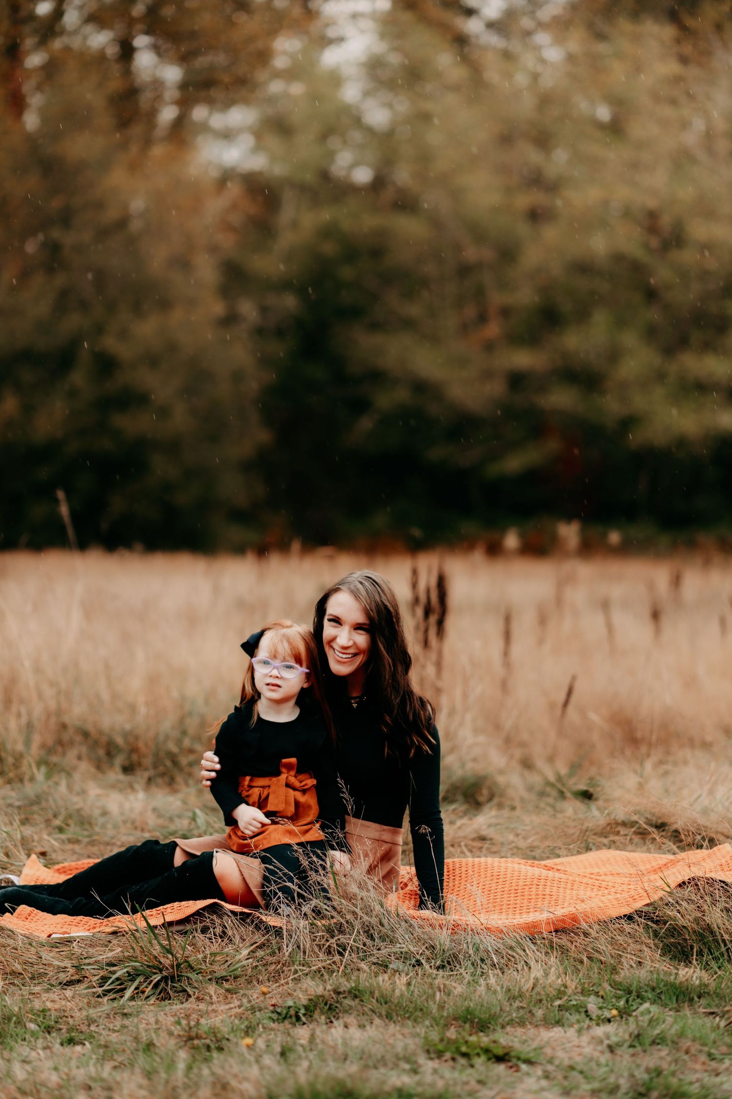 A woman and young child sit together on an orange blanket in an autumn field, surrounded by golden grass and trees during family photos.