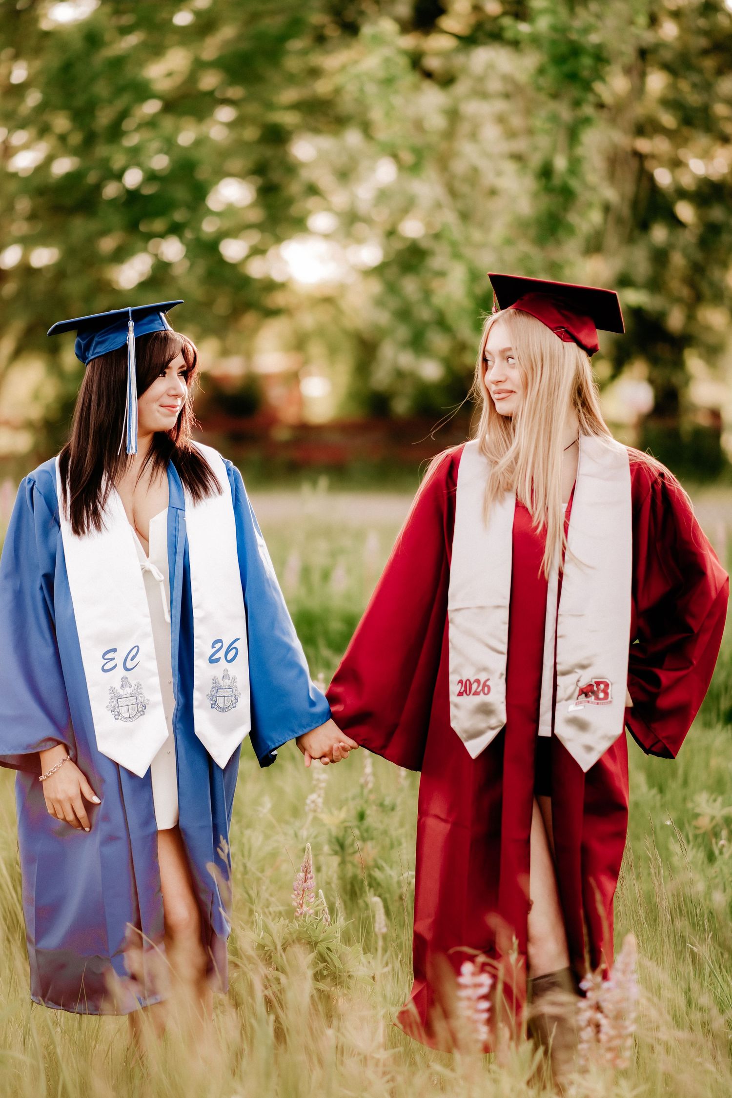Two graduates in blue and red caps and gowns hold hands walking through a green field during high school senior photos in Puyallup, Wa.