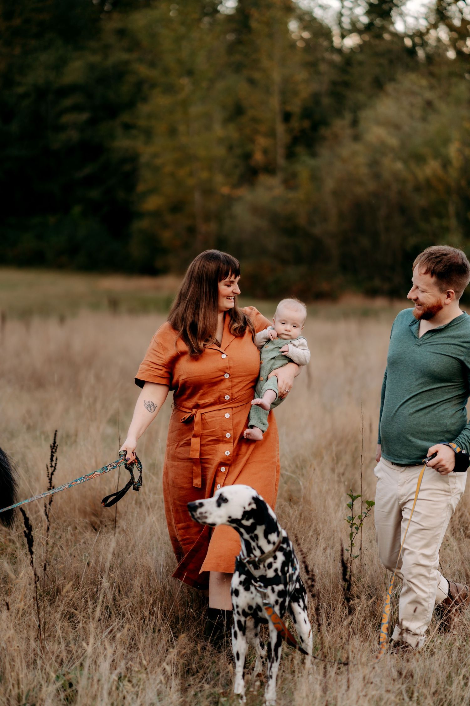 Family with baby and Dalmatian dog walking in autumn field, woman in orange dress, man in green shirt during family photos in Tacoma, WA.