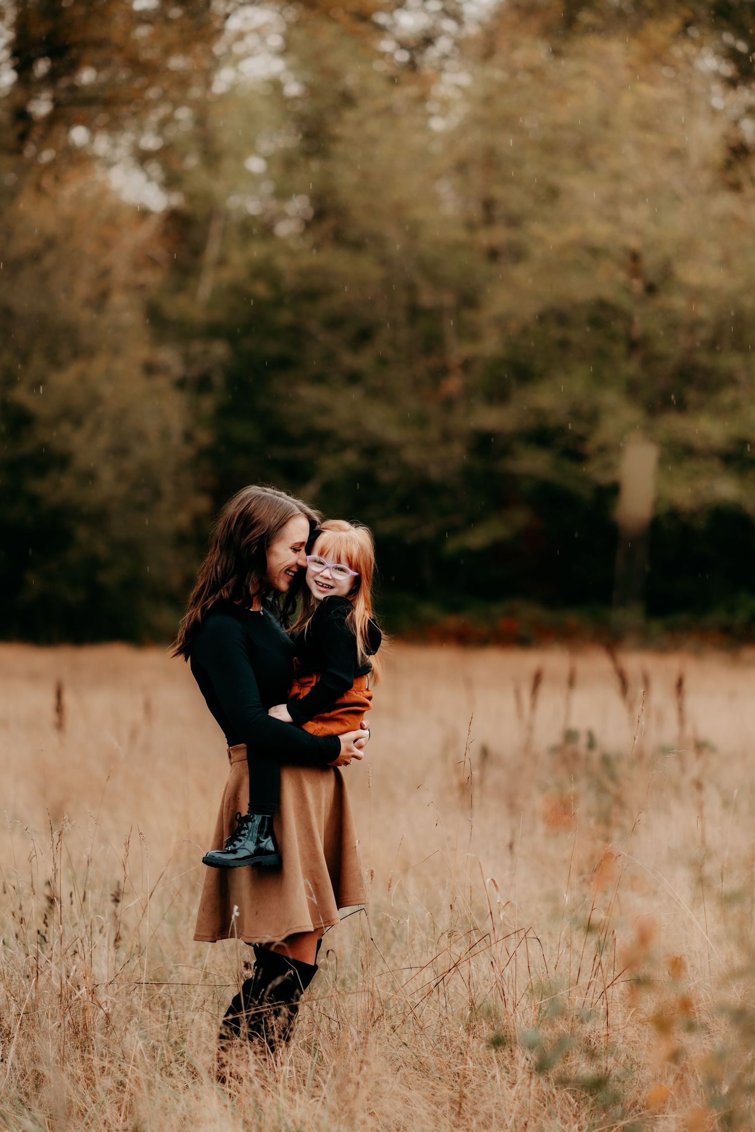 A mother holds her young child in a golden autumn field, both smiling warmly, surrounded by tall grass and fall foliage.