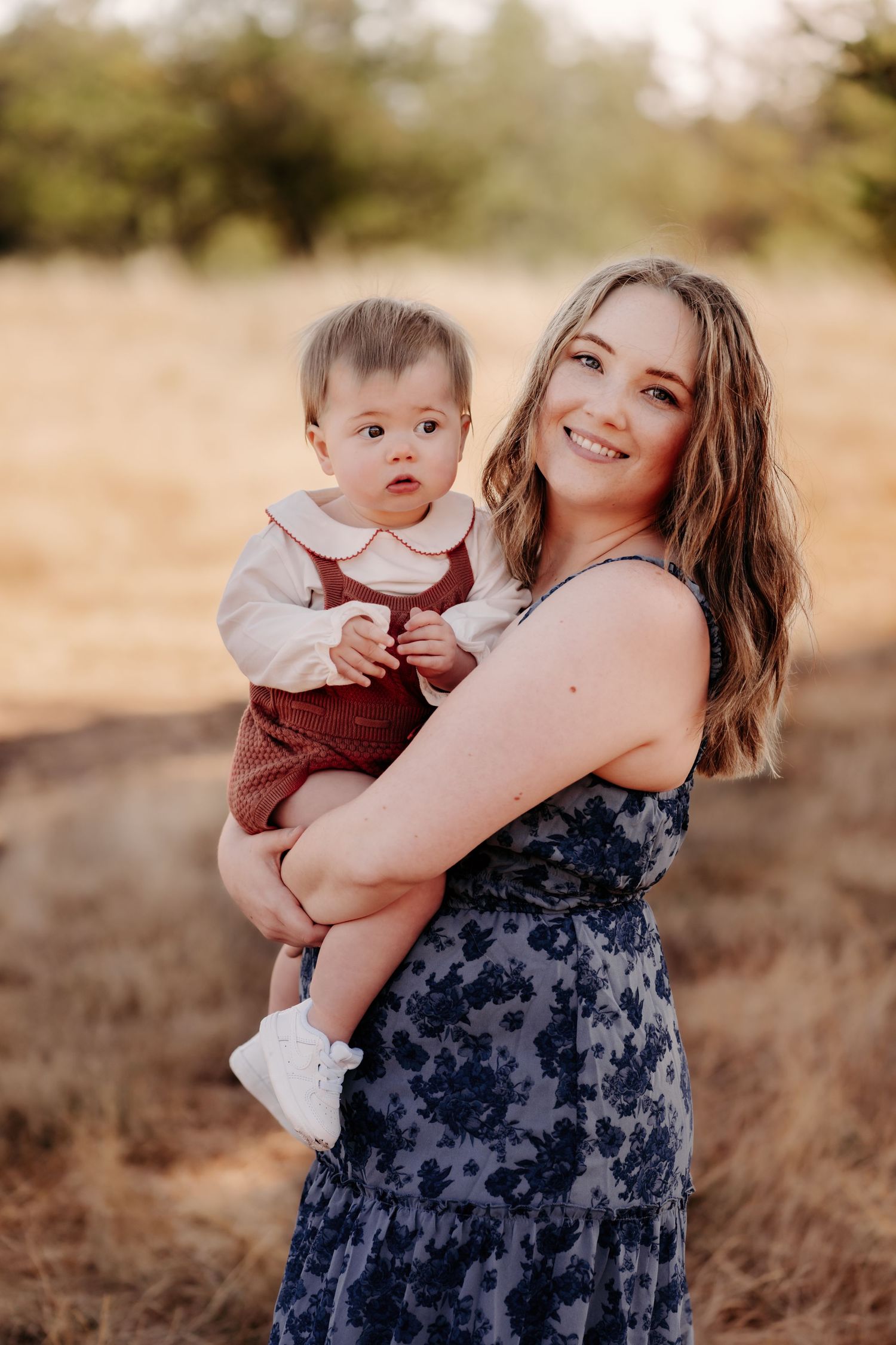 Smiling young woman in floral dress holding an adorable baby in red romper outdoors in golden field during family photo session in Tacoma.