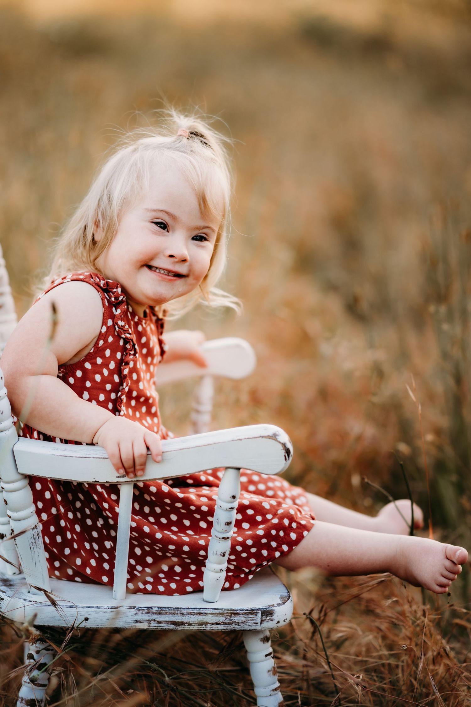 Smiling toddler girl with blonde hair in red polka dot dress sitting on white chair in golden field during family photo session in Tacoma.