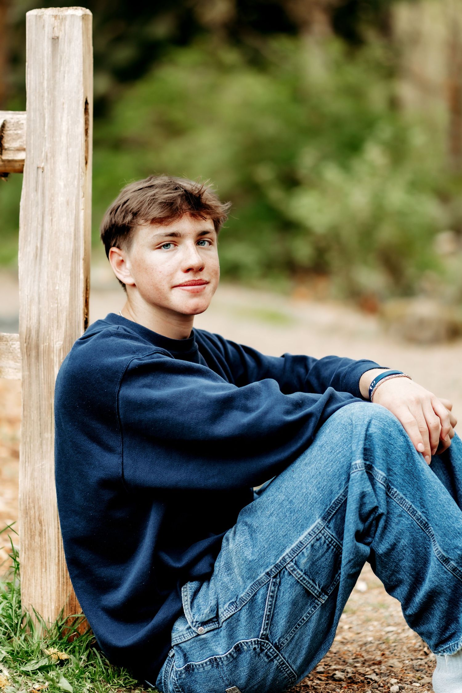 A teenage boy in a navy sweatshirt and jeans sits relaxed against a wooden post outdoors, smiling softly.