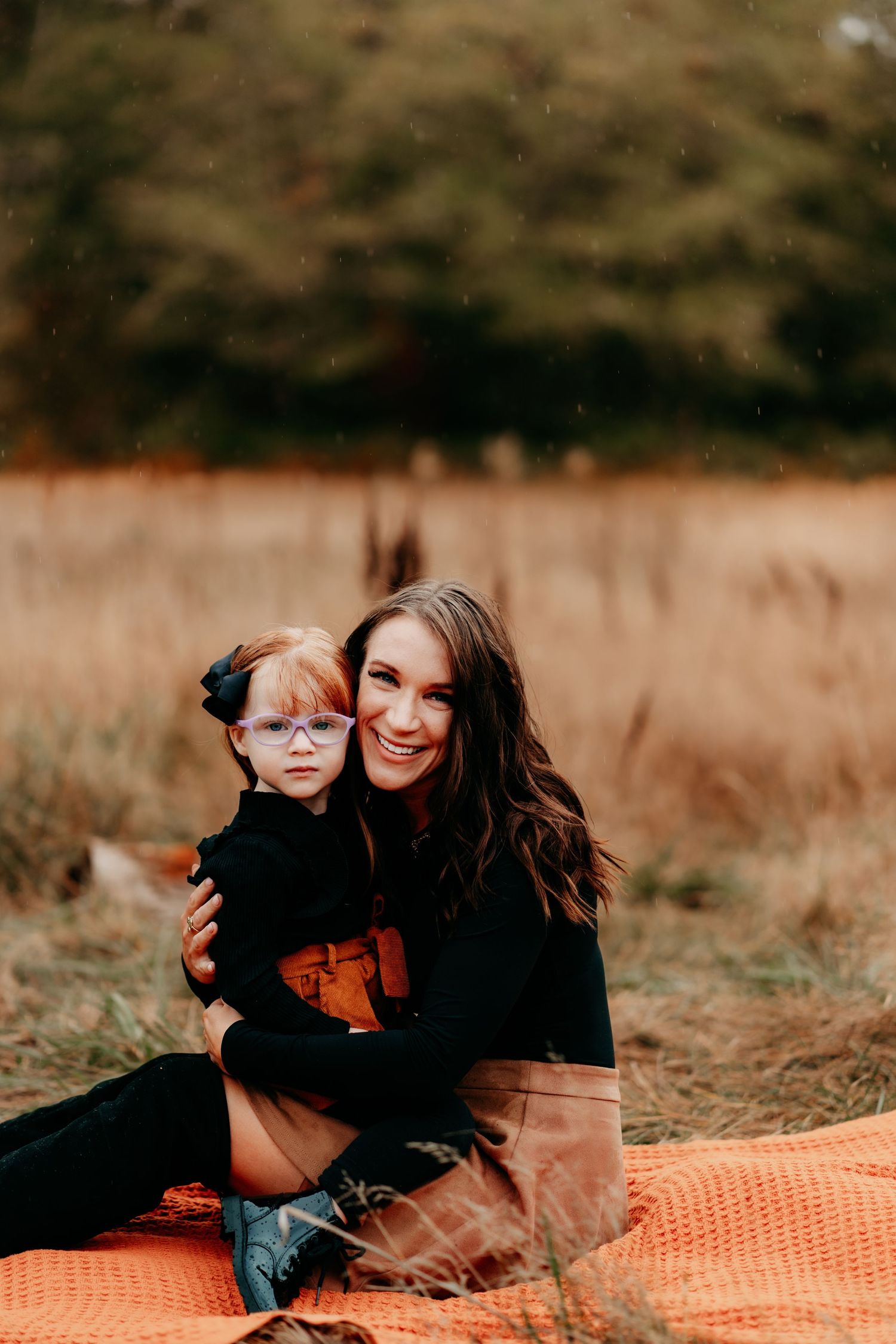 A smiling woman hugs a young girl with glasses on an orange blanket in an autumn field during family photos in Tacoma, Wa.