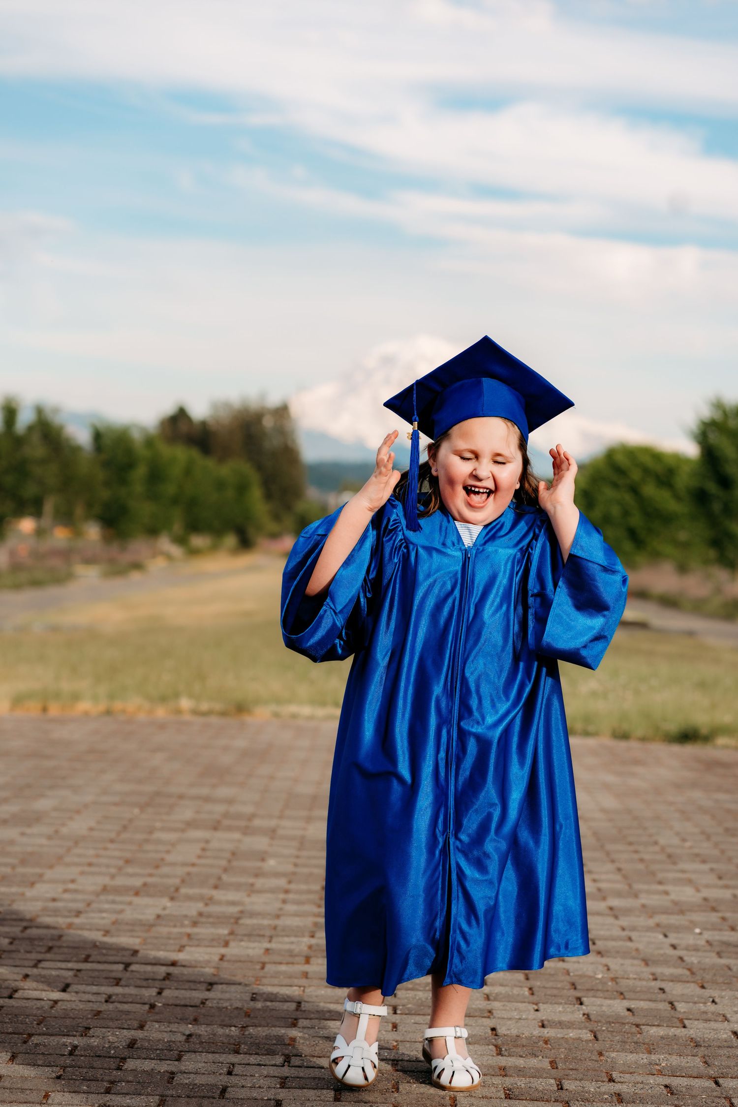Excited young girl in blue graduation cap and gown celebrating outdoors on a sunny day.
