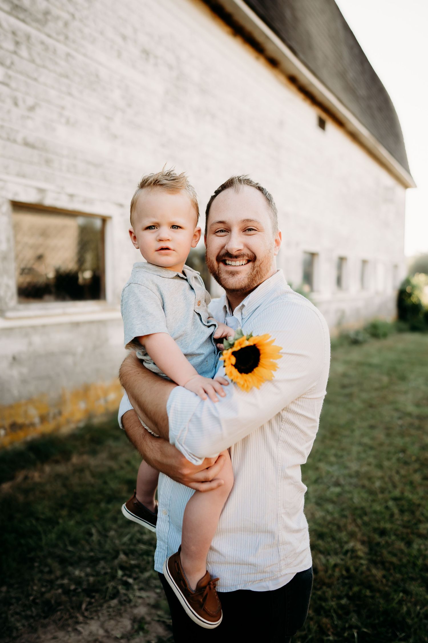 Smiling man holding toddler boy with sunflower outdoors near white barn during golden hour family photo session in Tacoma, WA.