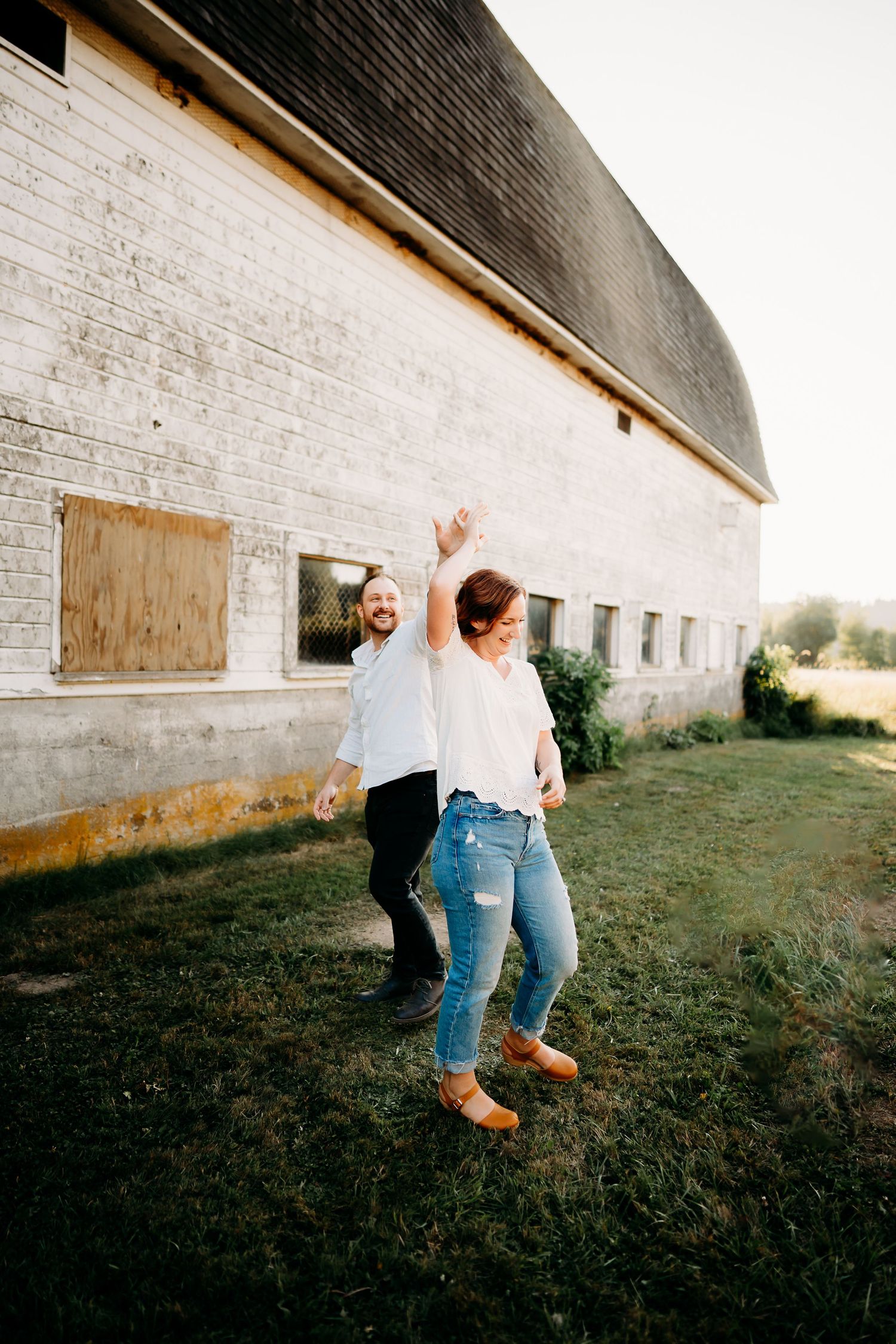 A joyful couple dancing and laughing outdoors beside a rustic white barn on a sunny day during golden hour family photos in Puyallup, Wa.