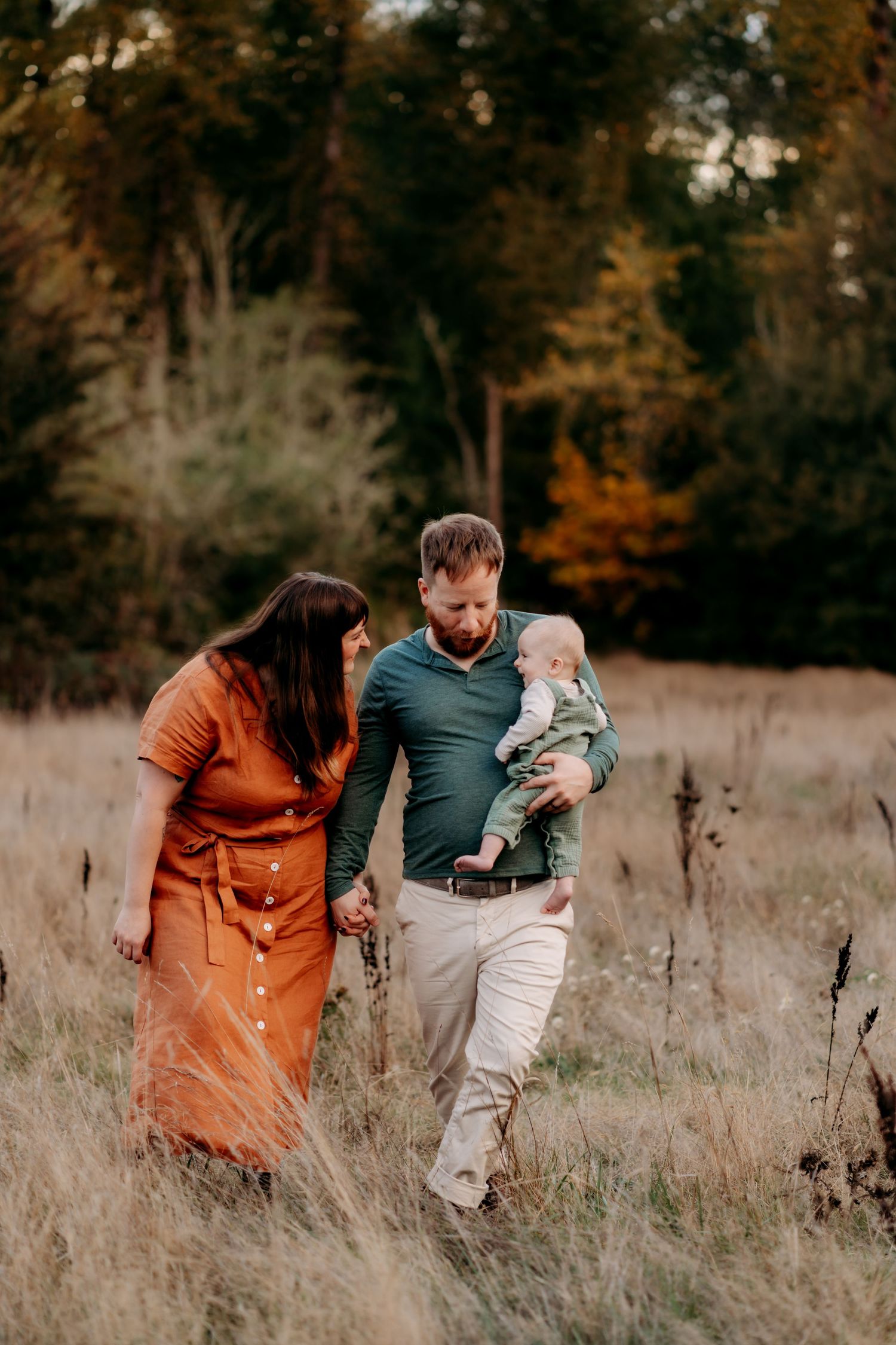 A family of three walks through a golden autumn field, parents holding hands while dad carries their baby at family photo session in Tacoma.