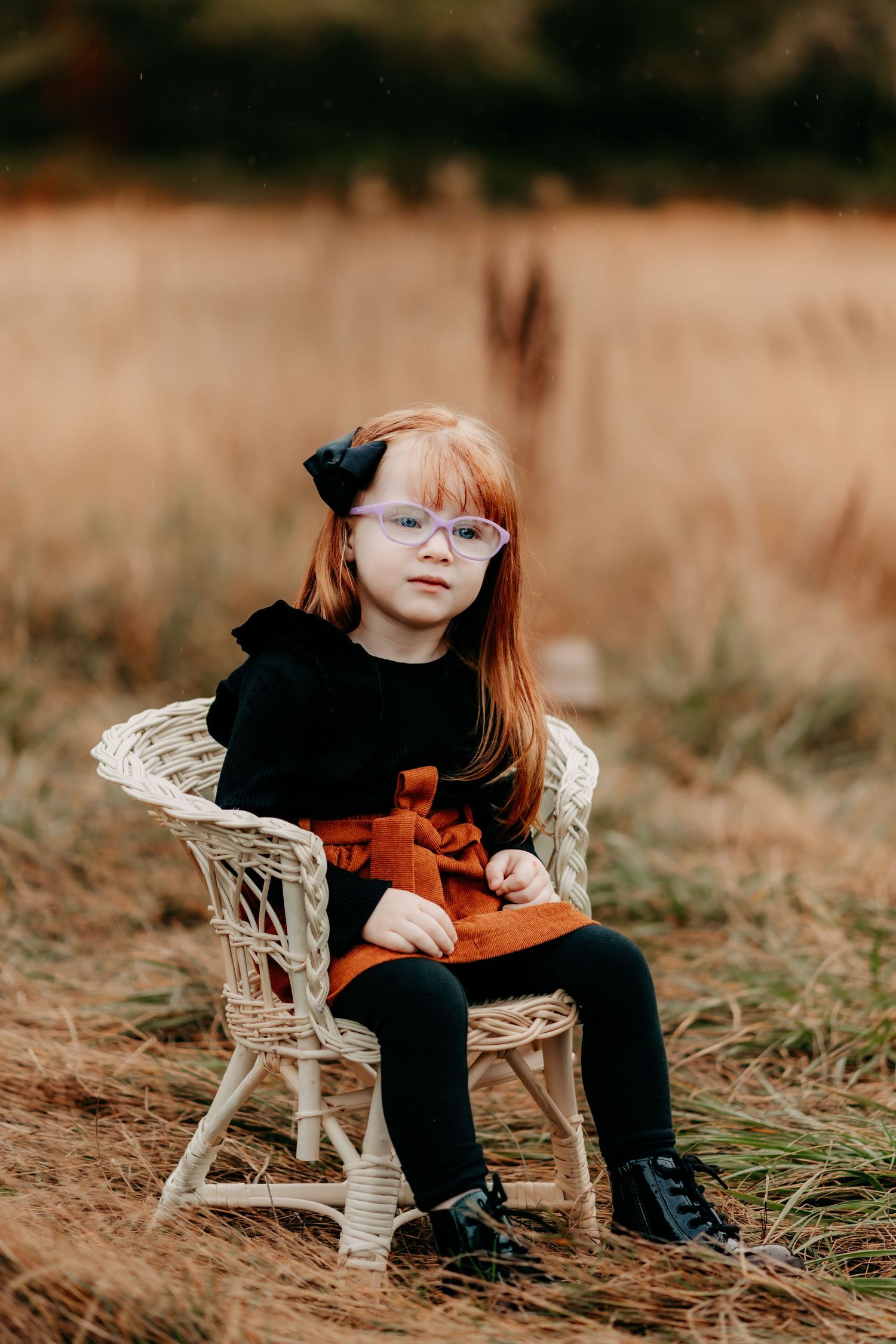 A young red-haired girl in glasses sits on a wicker chair in a golden field wearing a black top and rust skirt during fall photo session.