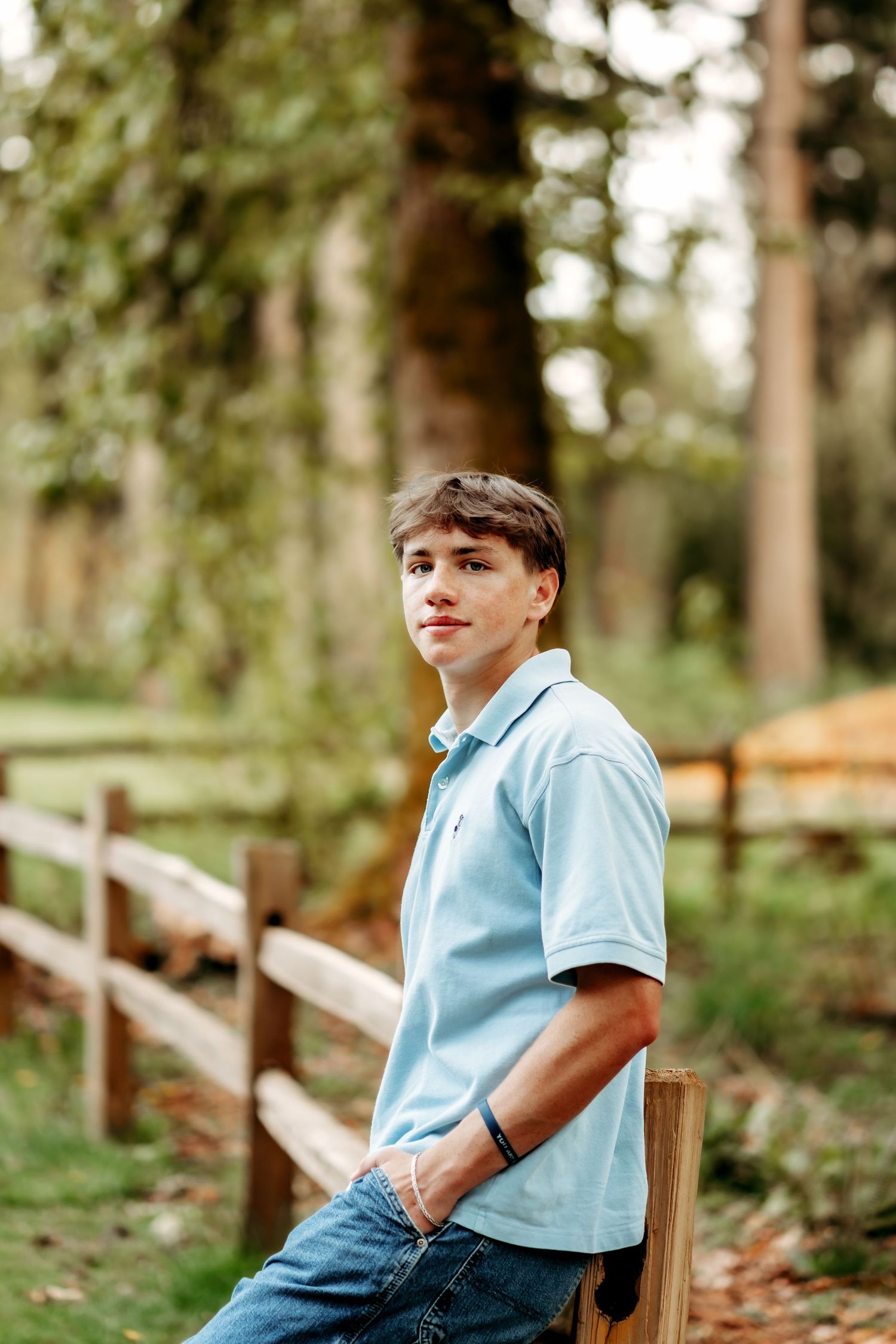 Teen boy in light blue polo shirt leans on wooden fence in lush green forest setting during high school senior photos in Puyallup, Wa.