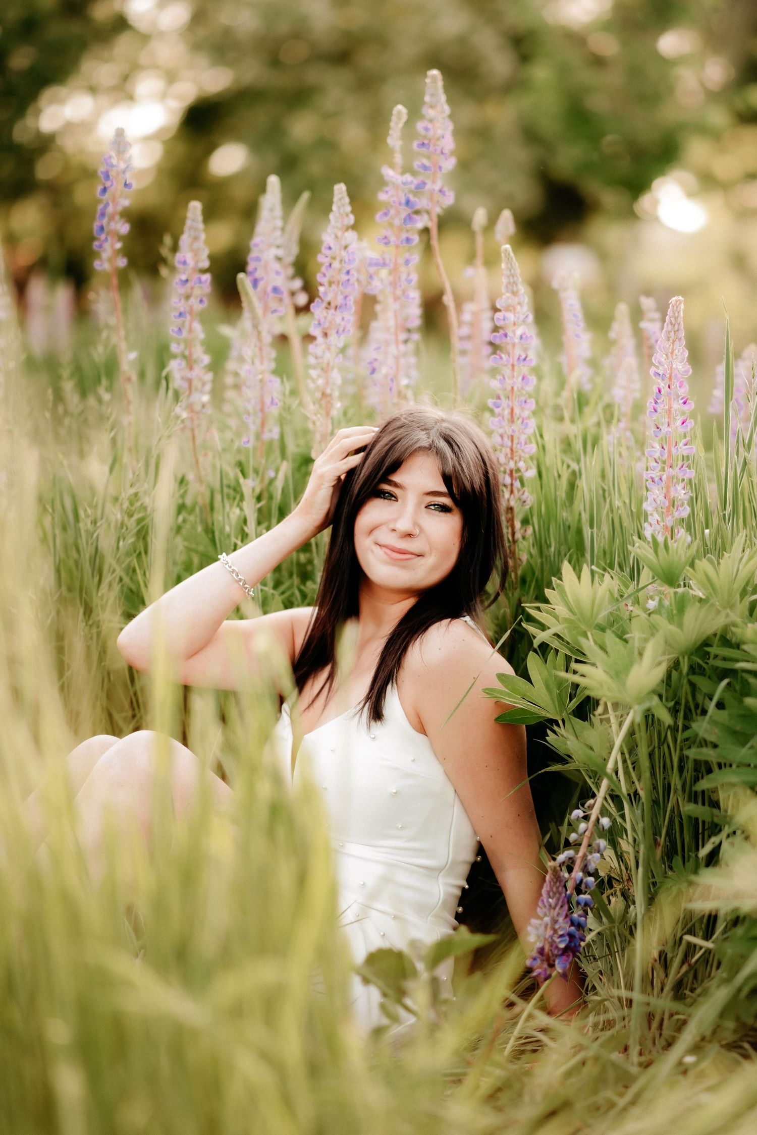 Young woman with dark hair sits smiling among tall purple lupine flowers in a lush green field wearing a white dress during senior photos.