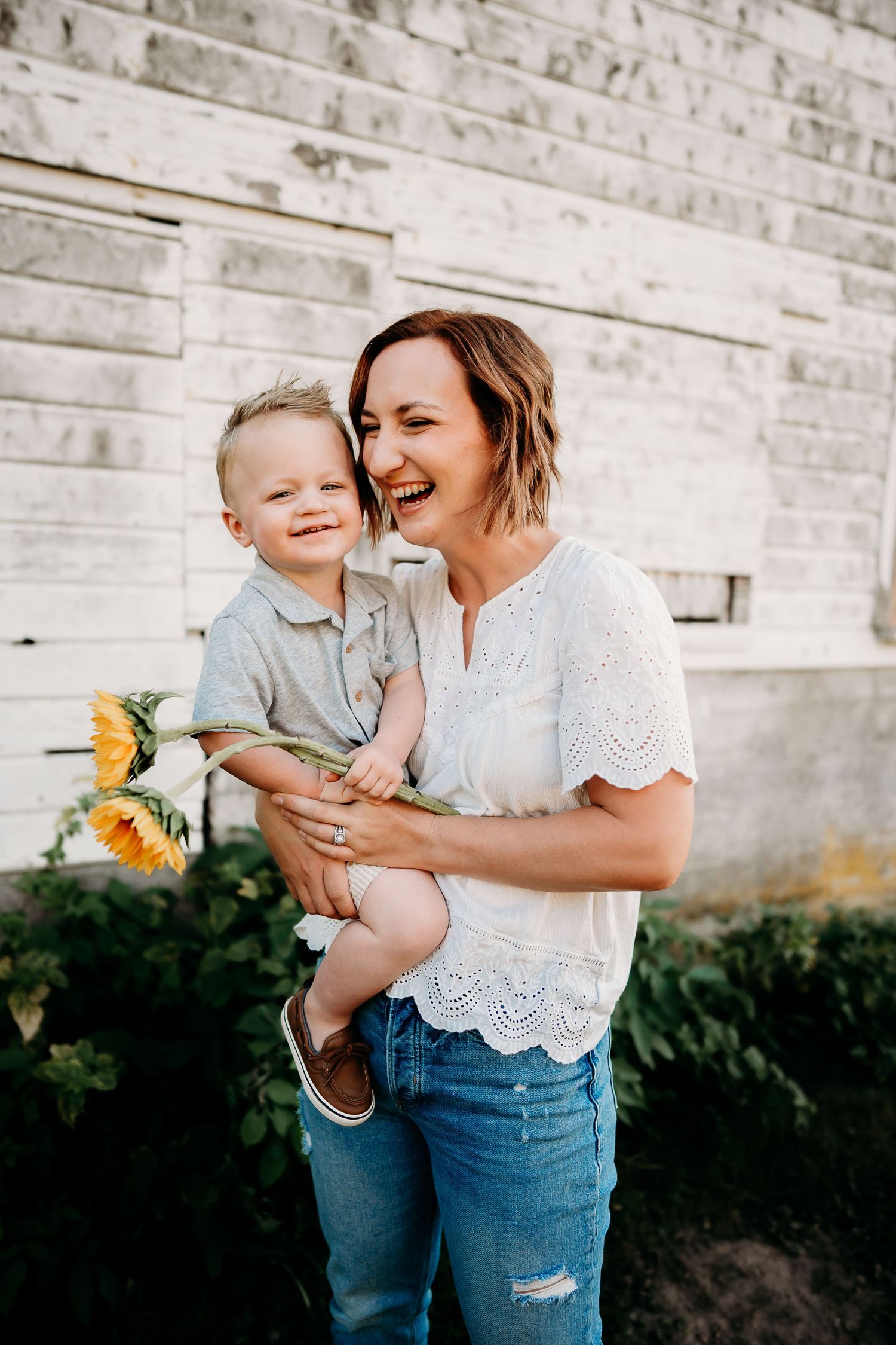 A laughing woman holds a young boy clutching sunflowers, standing before a white barn wall during family photos in Tacoma, Wa.