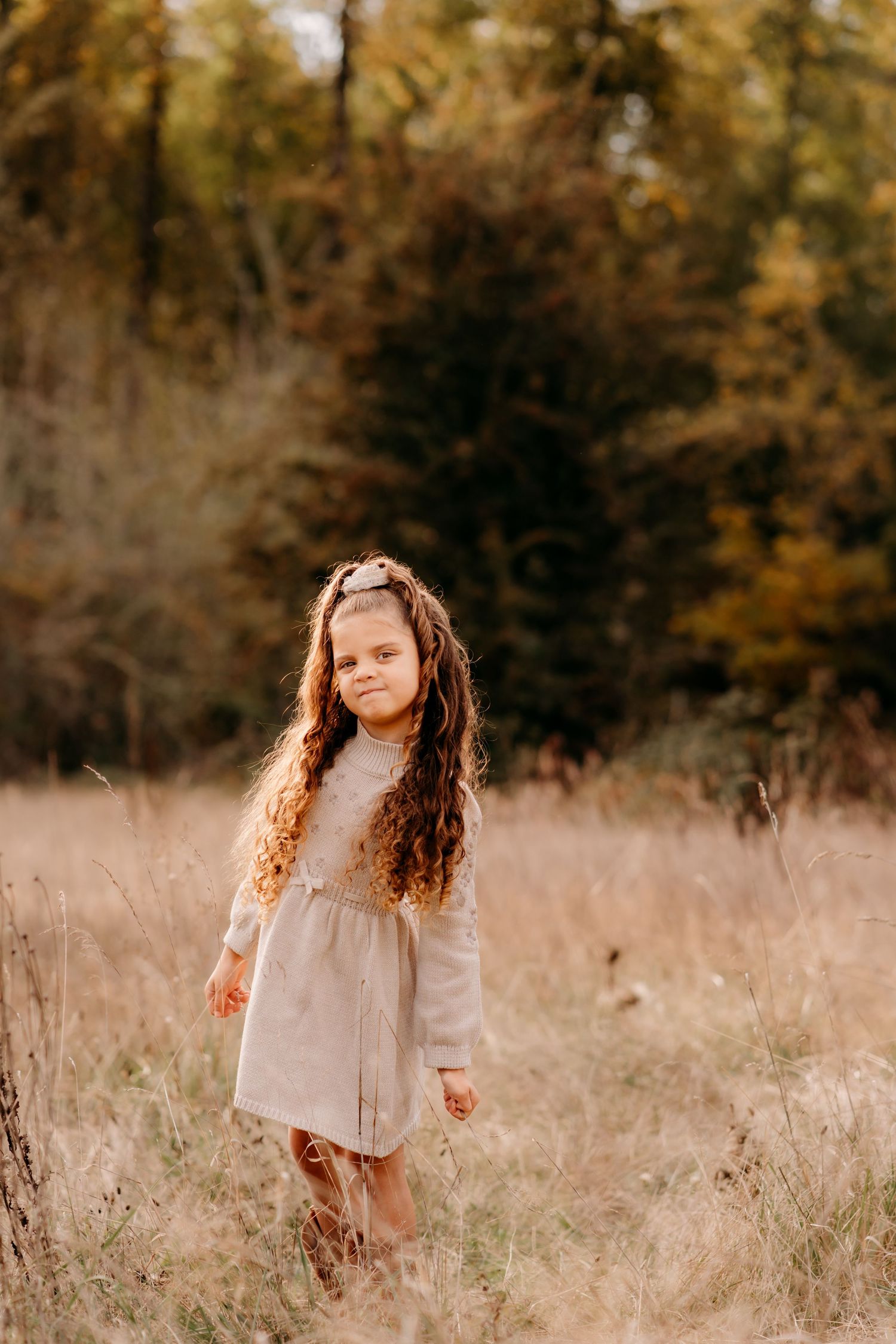 A young girl with long curly hair in a beige dress stands in a golden autumn field with colorful trees behind her during fall photo session.