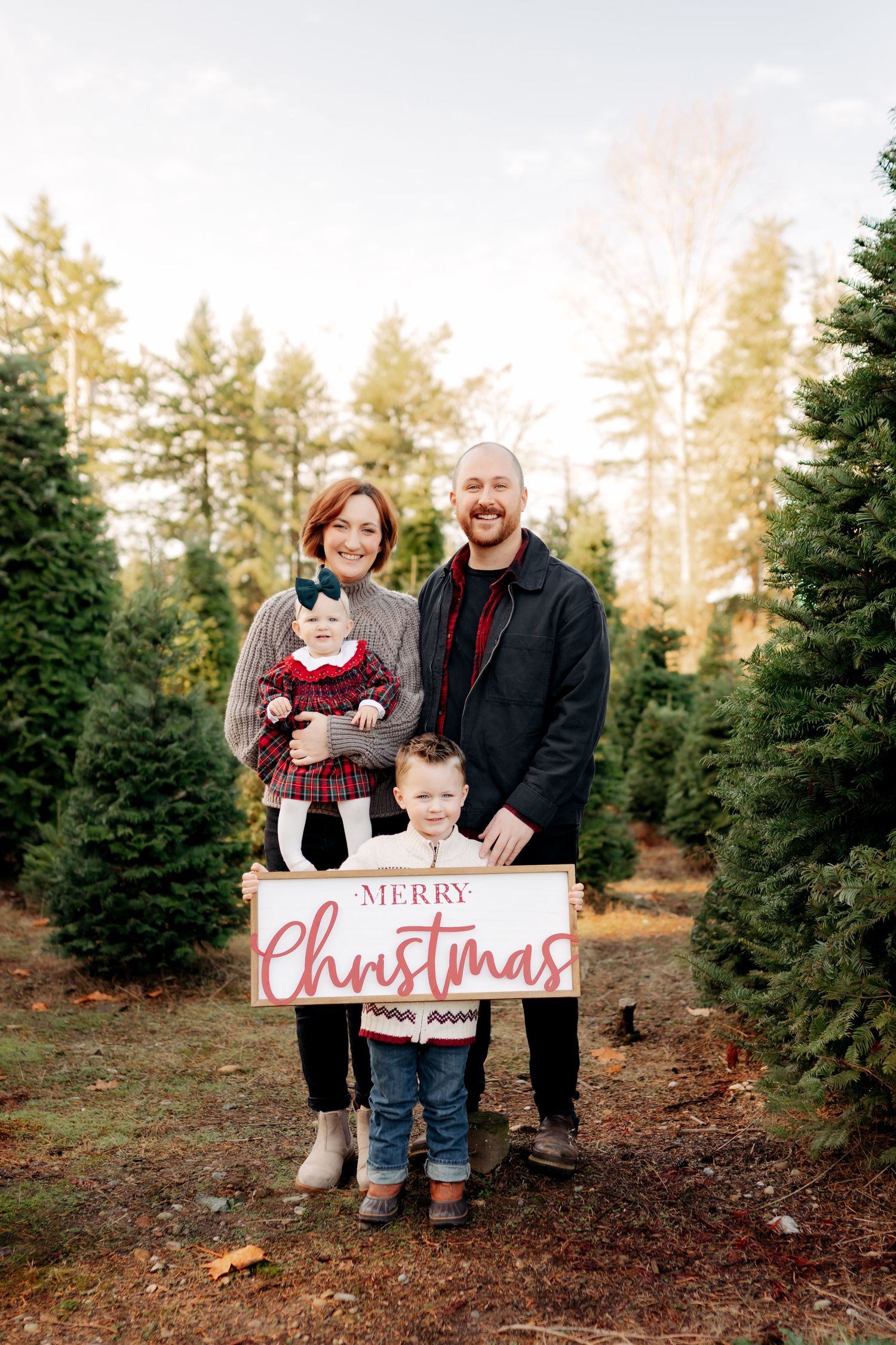 Family of four posing with Merry Christmas sign at Christmas tree farm, parents holding baby girl with toddler son in front.