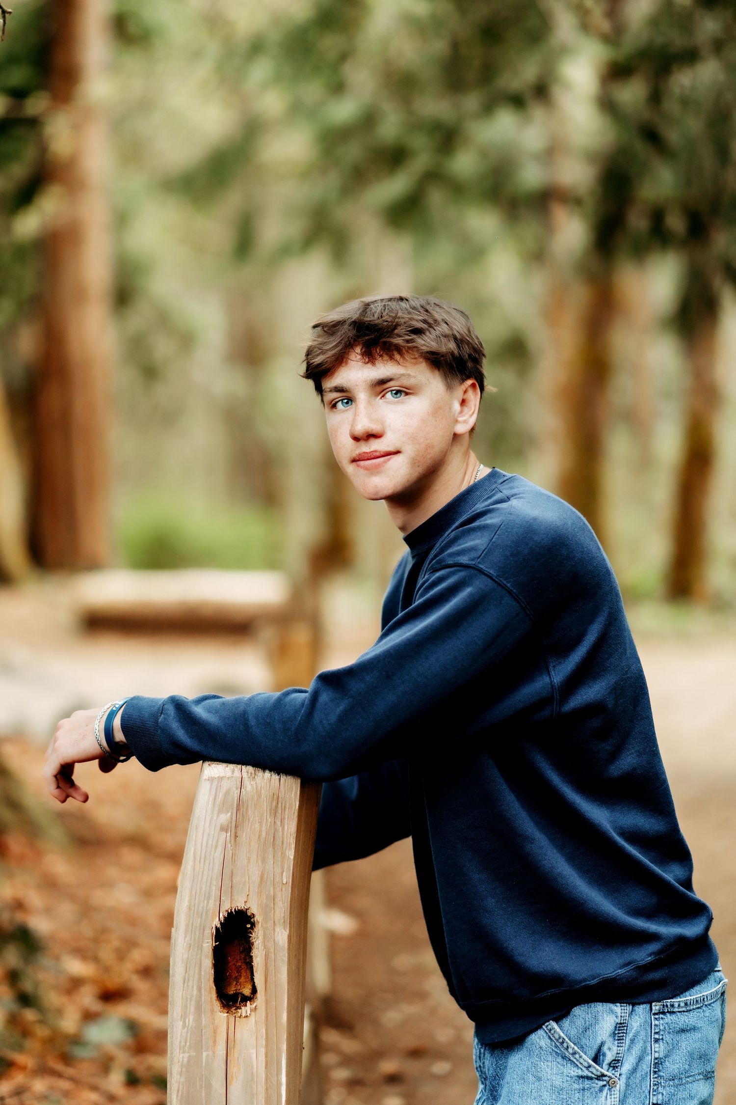 Young man in navy sweater and jeans leans on wooden post smiling in a lush forest setting for his high school senior photos in Puyallup, Wa.
