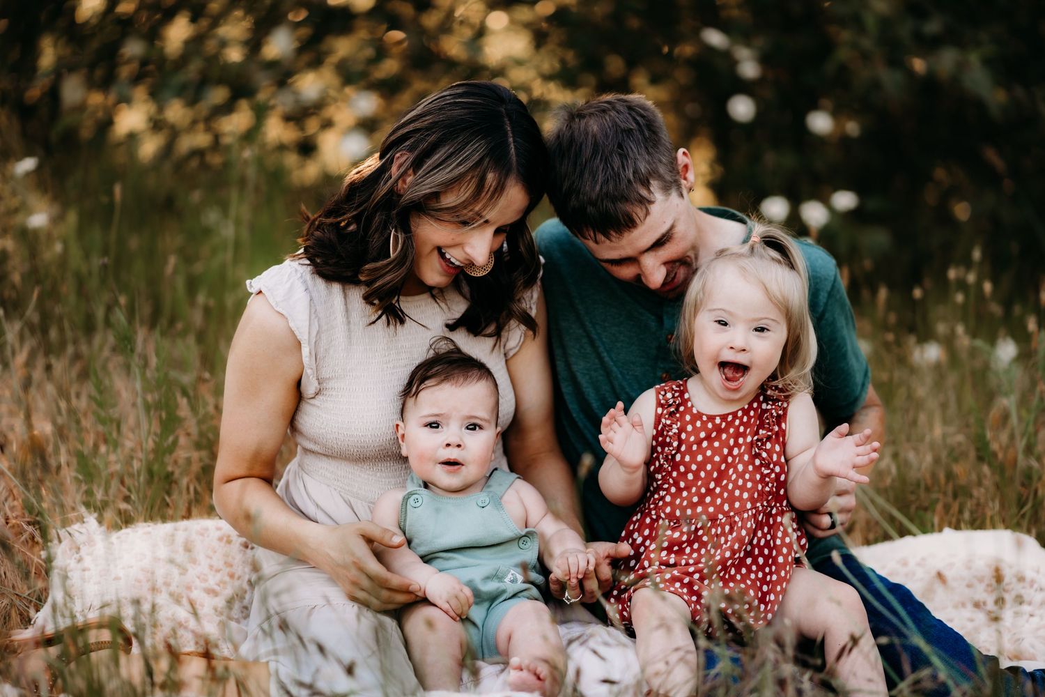 Happy family of four sitting outdoors in a meadow, parents smiling at two young toddlers on a sunny day, posing for family photos in Tacoma.