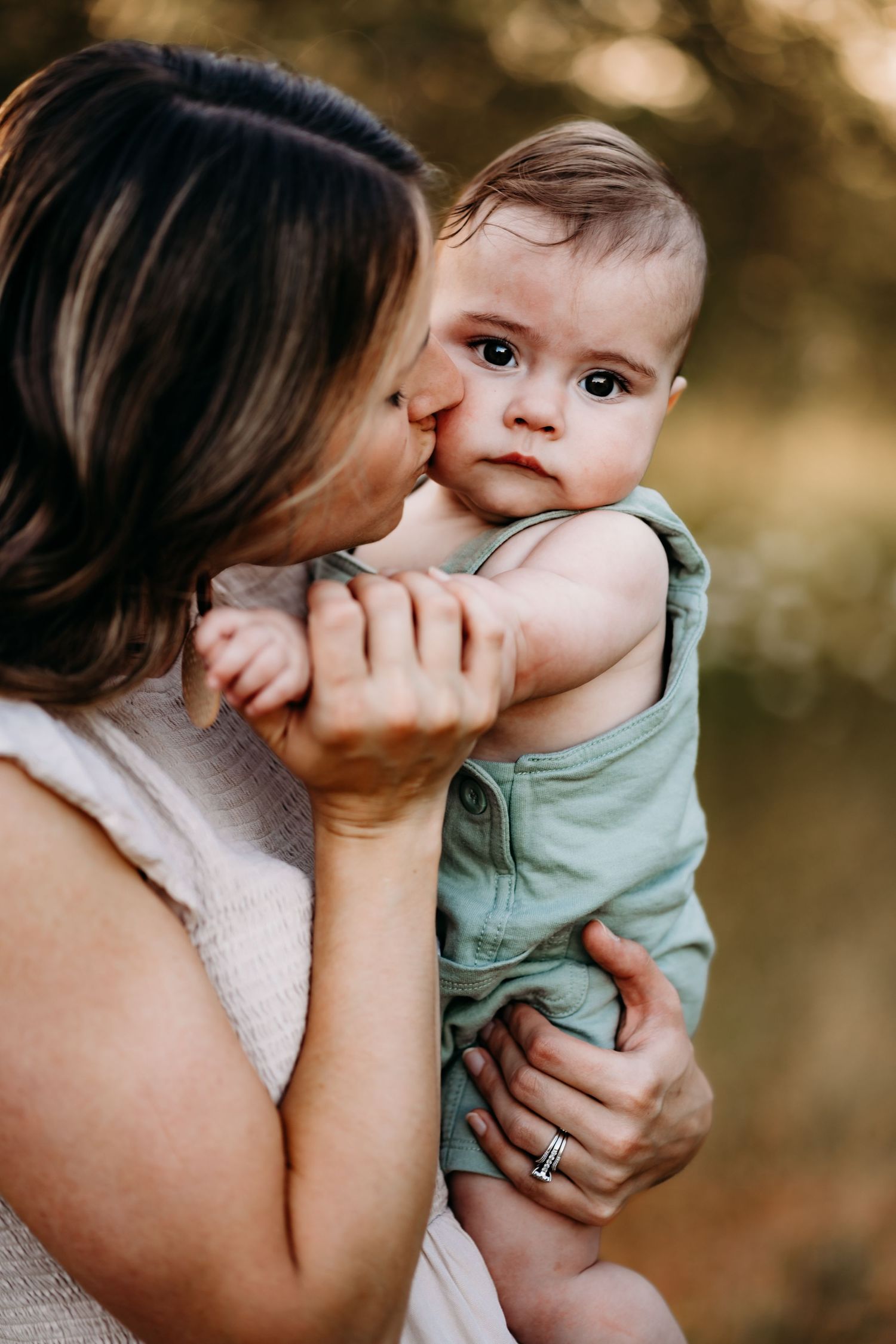 A mother tenderly kisses her baby's cheek while holding the infant dressed in a green romper during golden hour family photos in Puyallup.