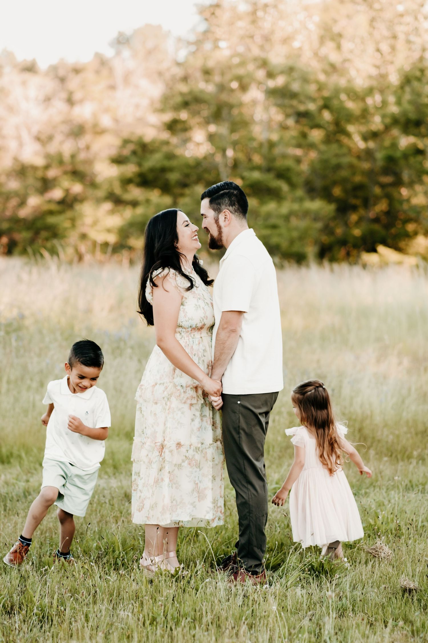 A family of four shares a tender moment in a sunlit meadow, parents holding hands while children play around them at their photo session.