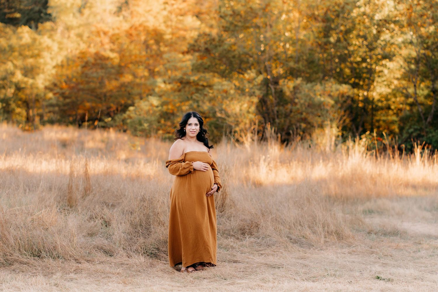 Pregnant woman stands in golden autumn field at sunset creating a warm and dreamy atmosphere for her maternity photo session in Tacoma.