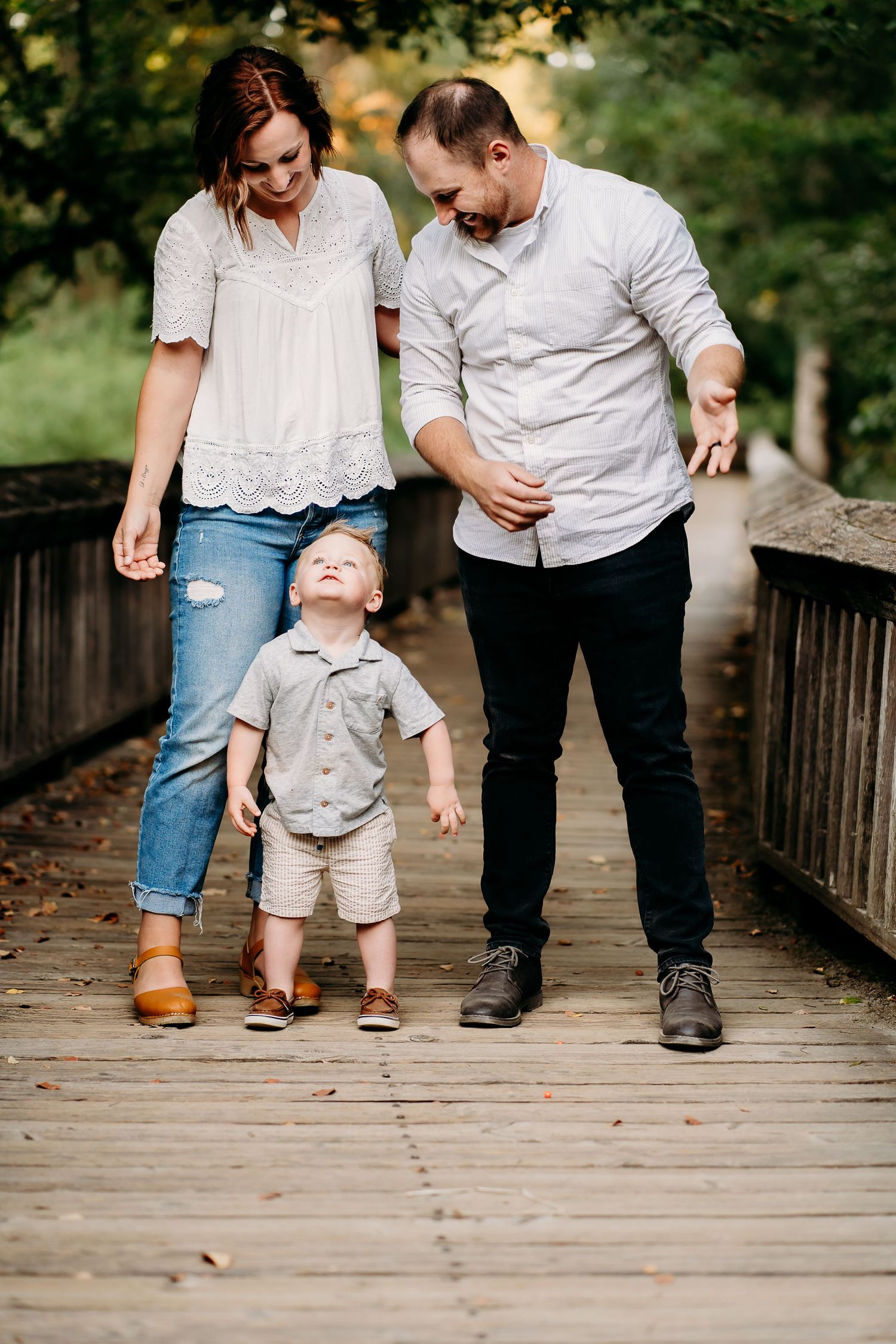 A young family with a toddler boy walking on a wooden bridge surrounded by lush green trees during golden hour family photos in Puyallup, Wa