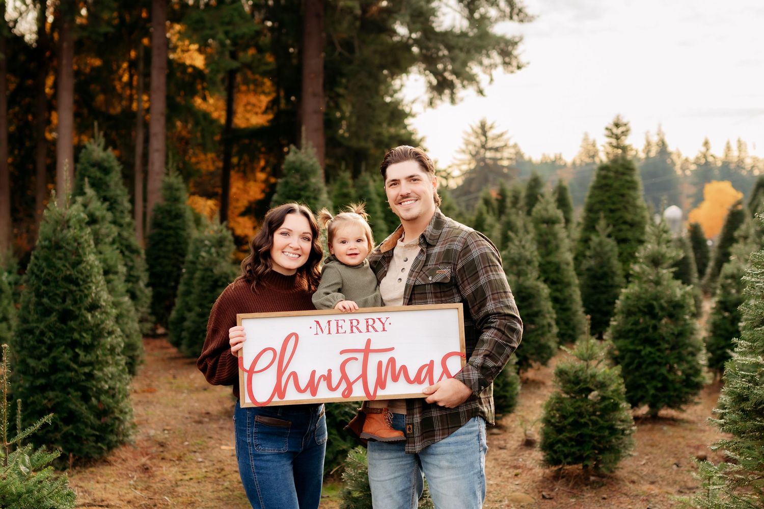 Family holding Merry Christmas sign at tree farm with evergreen trees in background during golden hour Christmas photo session in Tacoma.