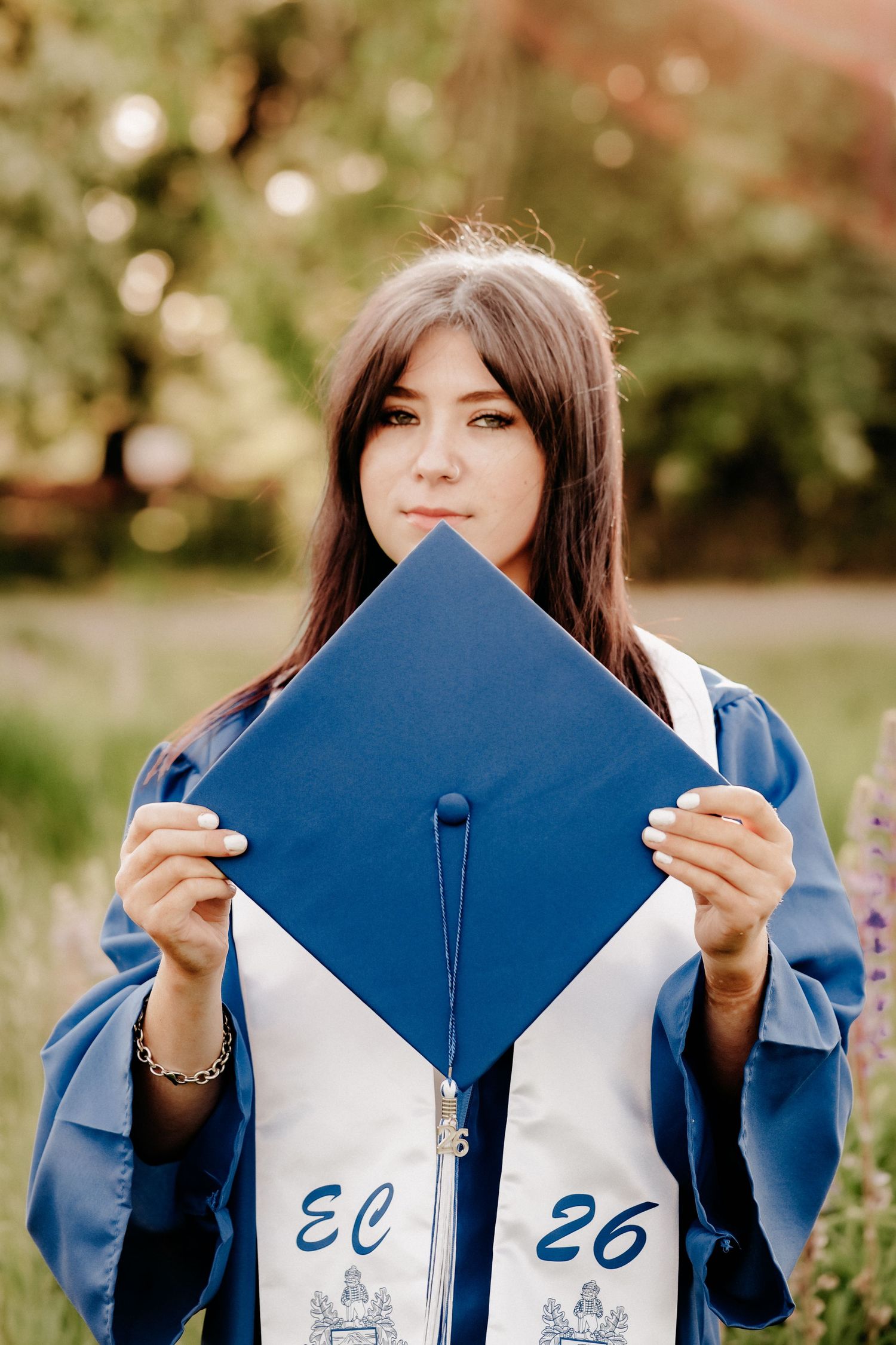 A young woman in blue graduation gown holds her mortarboard cap, with a stole reading EC 26, outdoors.