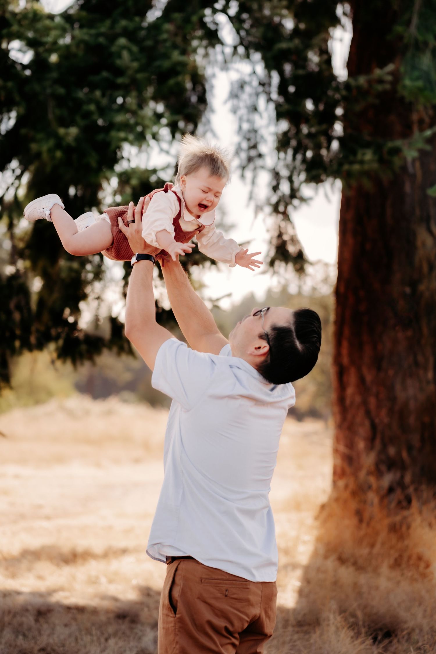 Father joyfully lifting laughing baby girl in the air outdoors in a sunny park setting during family photos in Puyallup, Wa.