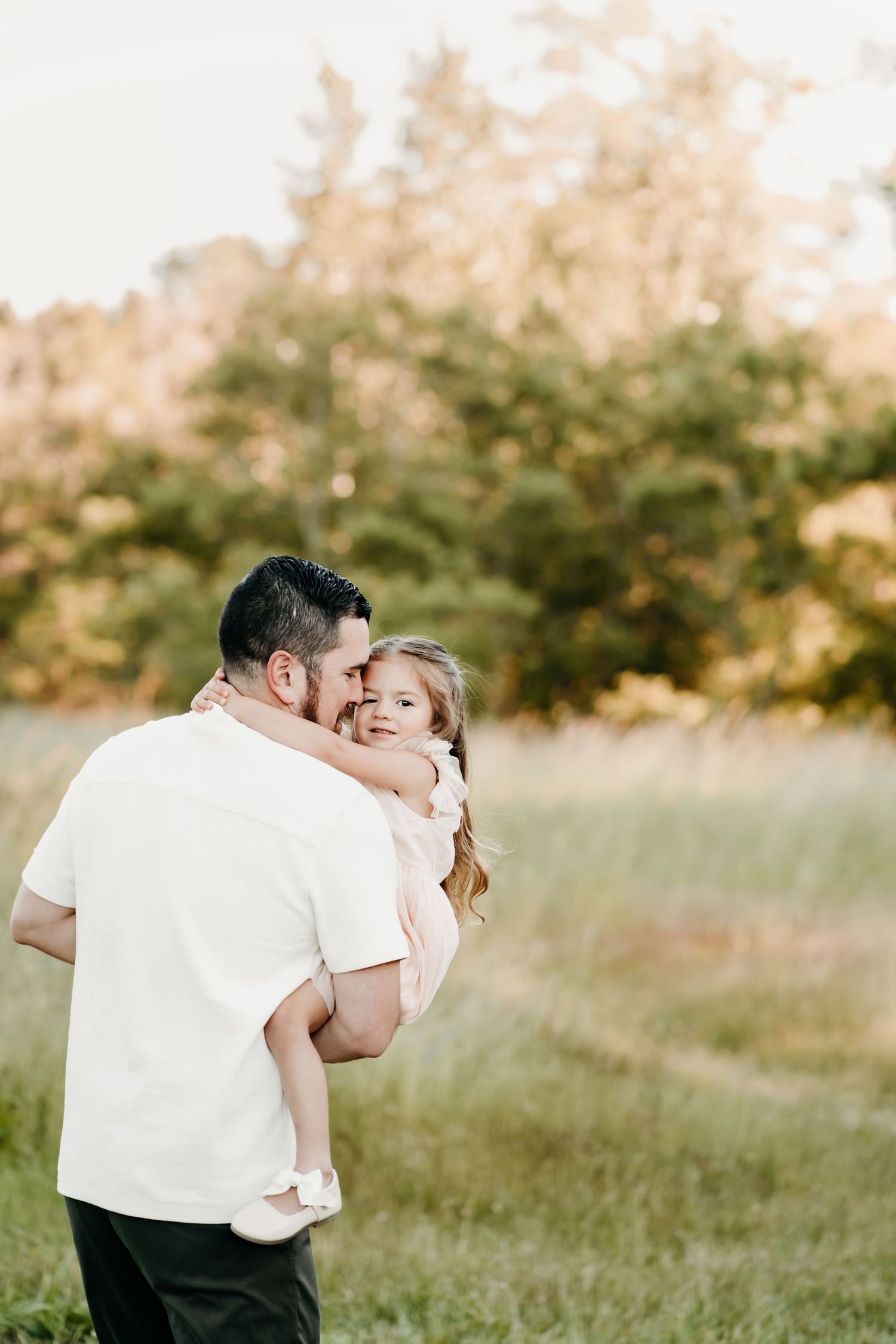 A father holds his young daughter in a sunlit field, sharing a tender moment with trees in the background during family photos in Tacoma, WA
