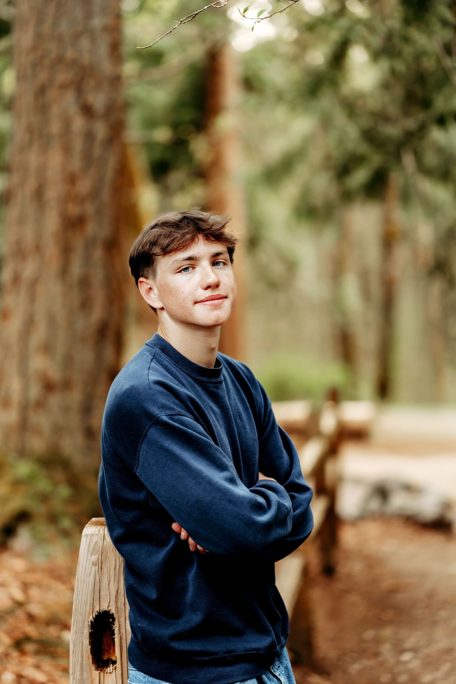 Young man in navy sweatshirt with arms crossed, smiling in a lush forest setting with tall trees during senior photo session in Tacoma, Wa.