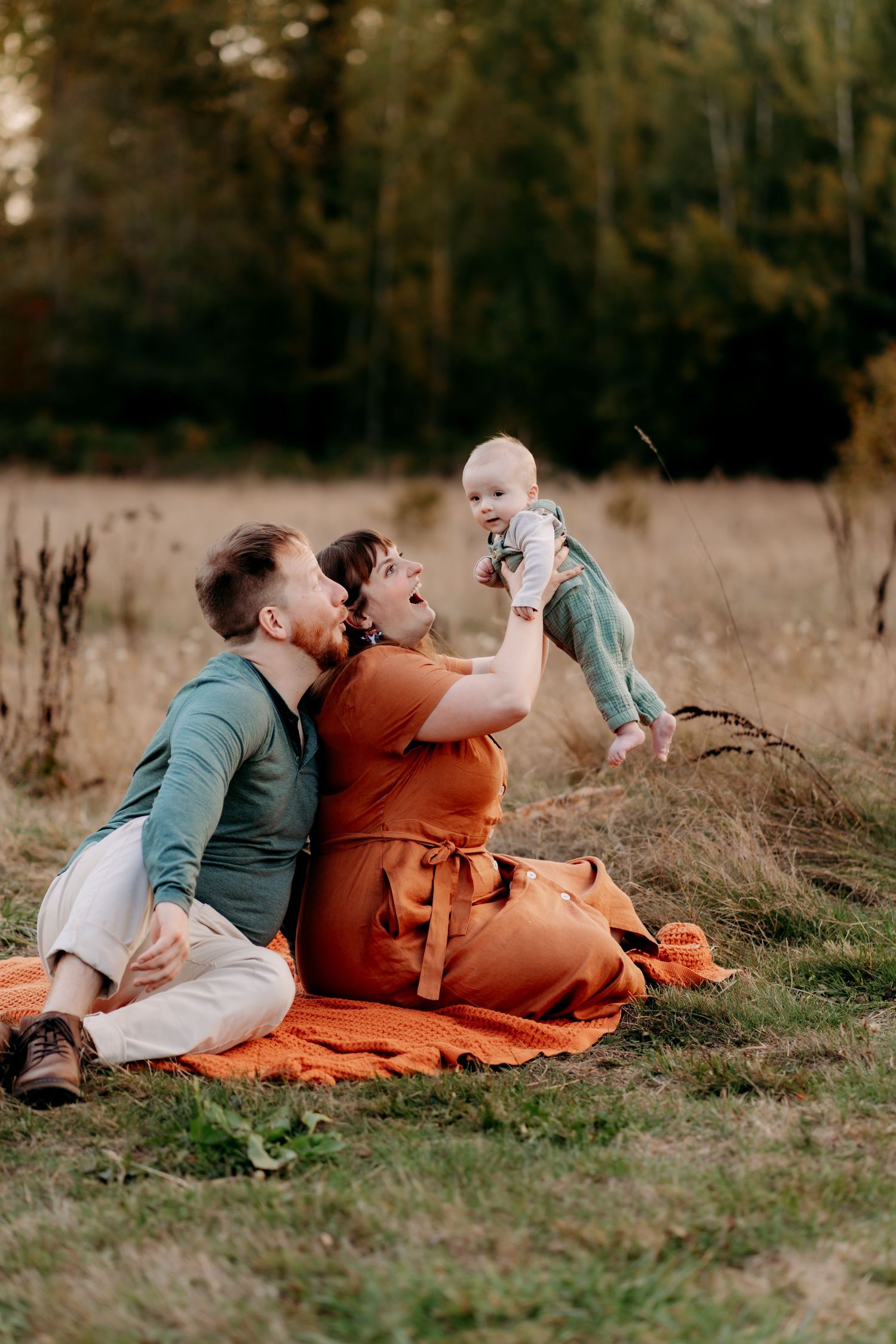 Family sitting on orange blanket in field, parents joyfully lifting smiling baby outdoors during golden hour photo session in Puyallup.