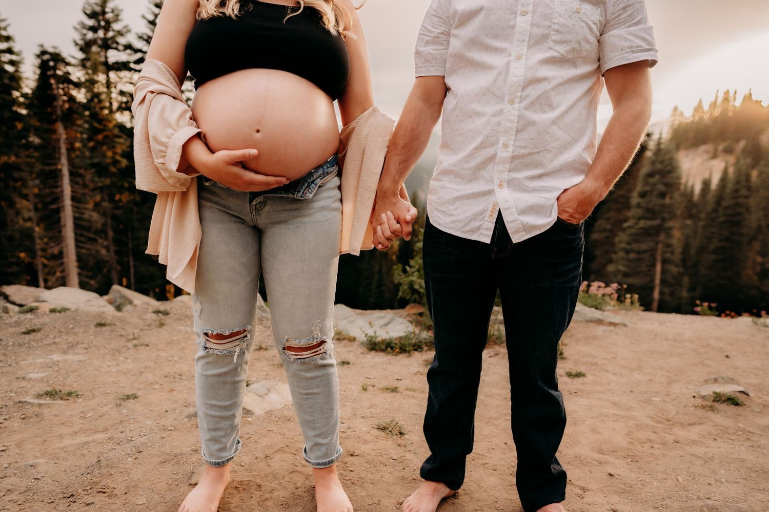 Husband and wife stand hand in hand, side by side, mountain side for maternity photos. Mom wears unbuttoned jeans with a bra leaving her bea