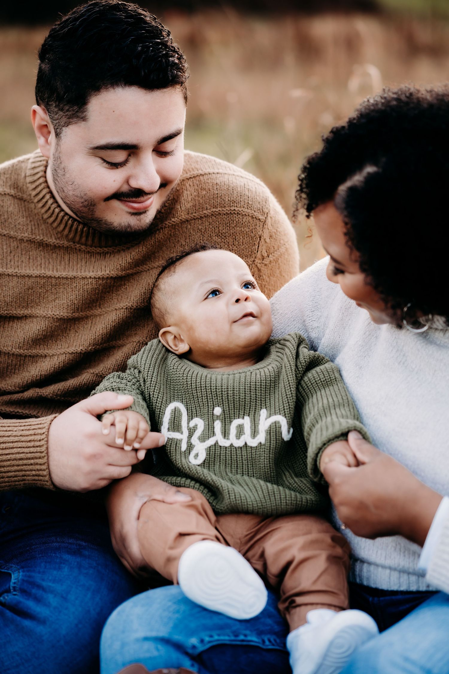 A baby wearing a green sweater sits between two adults in a warm outdoor family portrait.