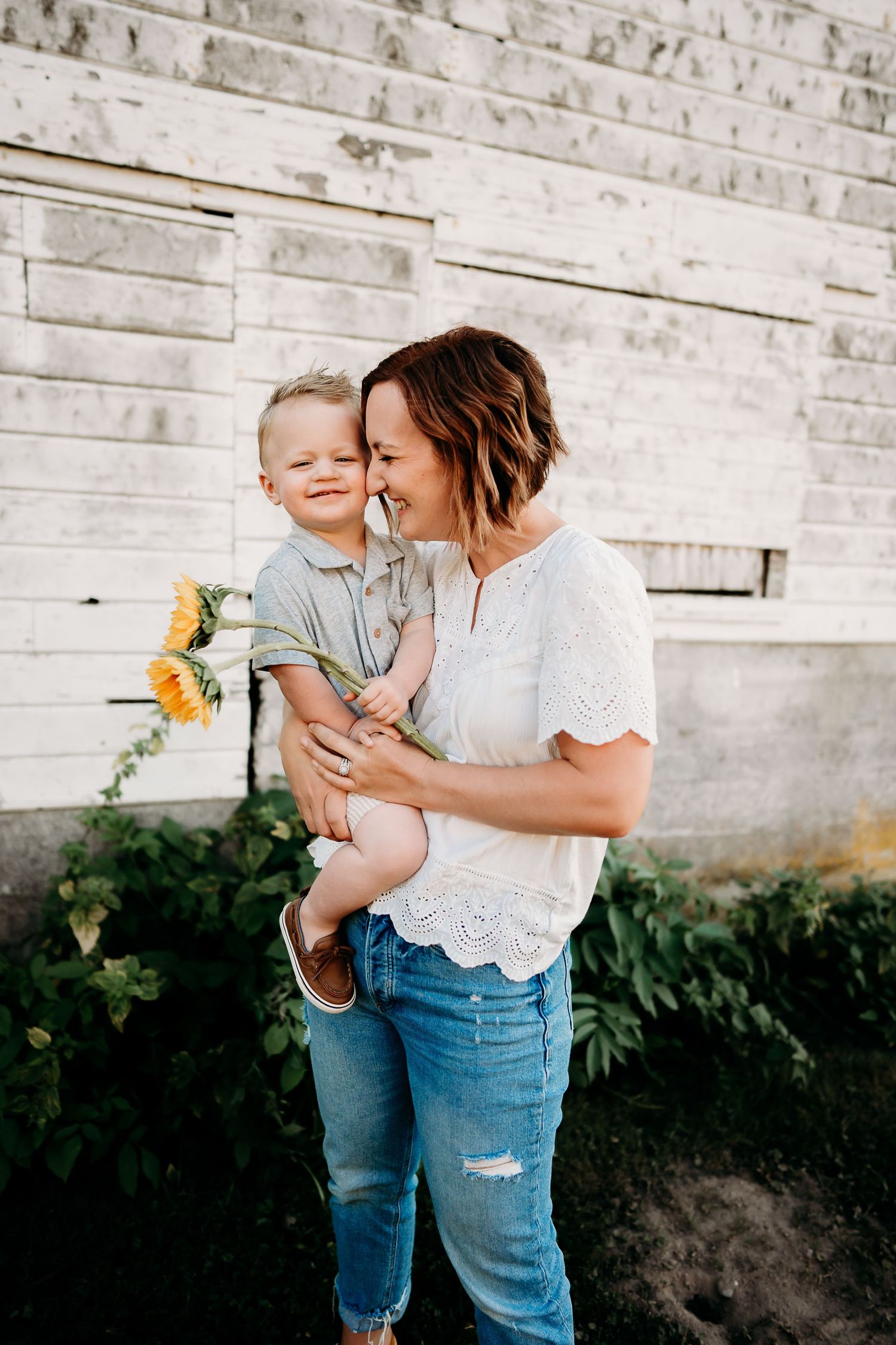 A laughing woman holds a young boy clutching sunflowers, standing before a white barn wall during family photos in Tacoma, Wa.