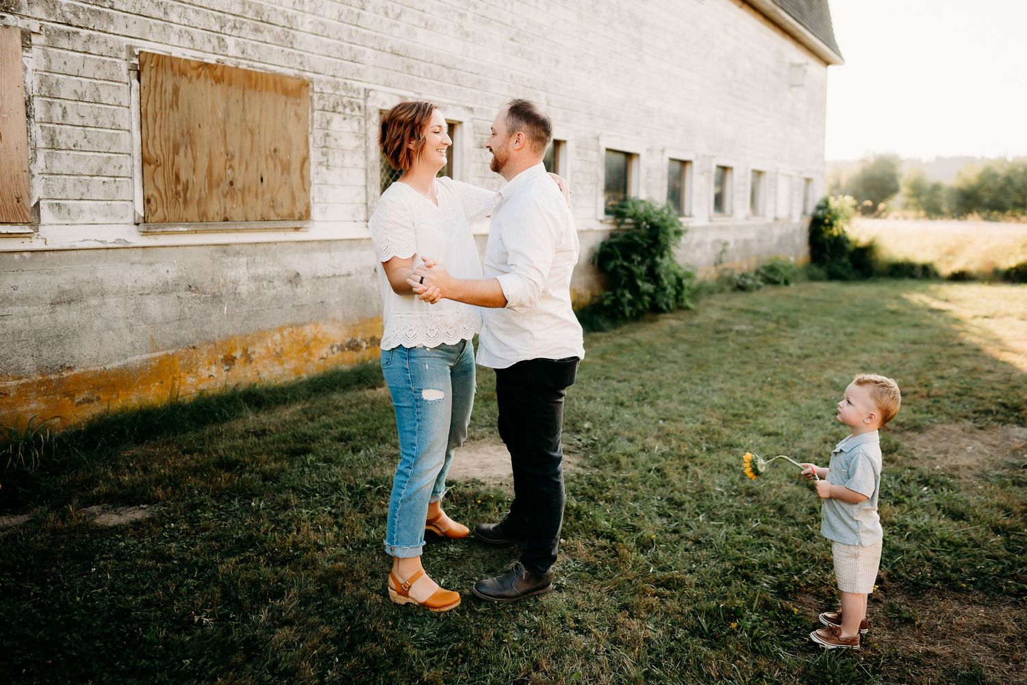 A young family with a toddler poses near a rustic barn in golden hour light for family photo session in Tacoma, Wa.