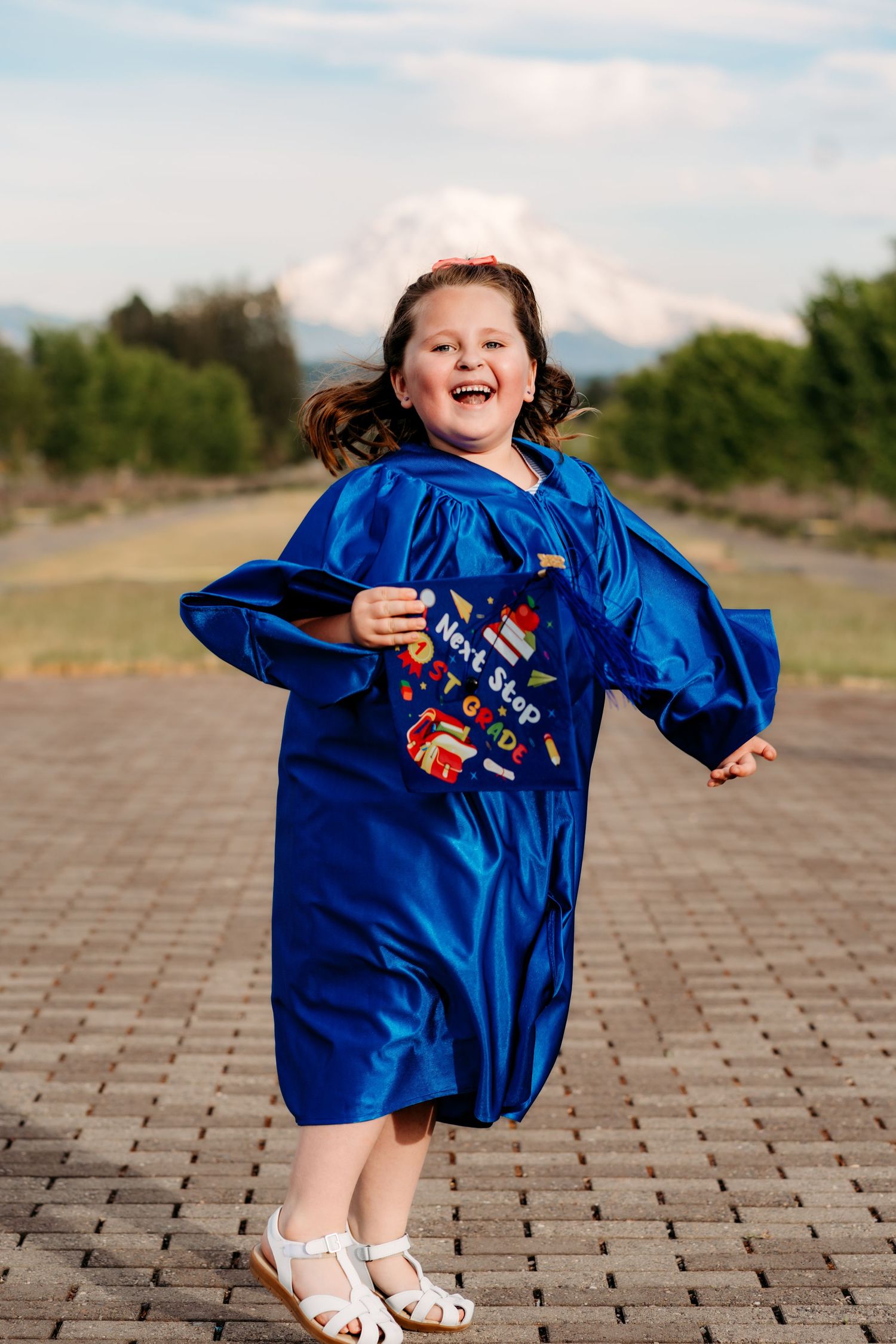 A joyful child wearing a bright blue graduation gown jumps and spins on a brick pathway with mountains in the background.