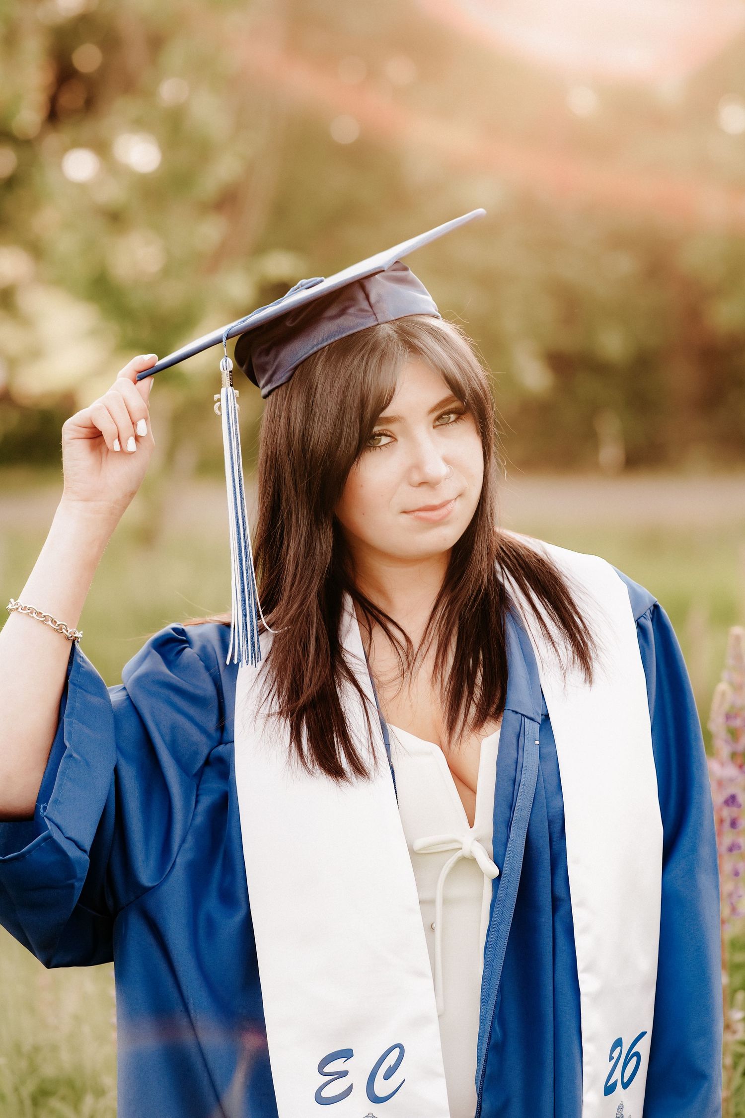 Young woman in blue graduation cap and gown holding tassel outdoors, wearing white stole with EC and 26 embroidered.