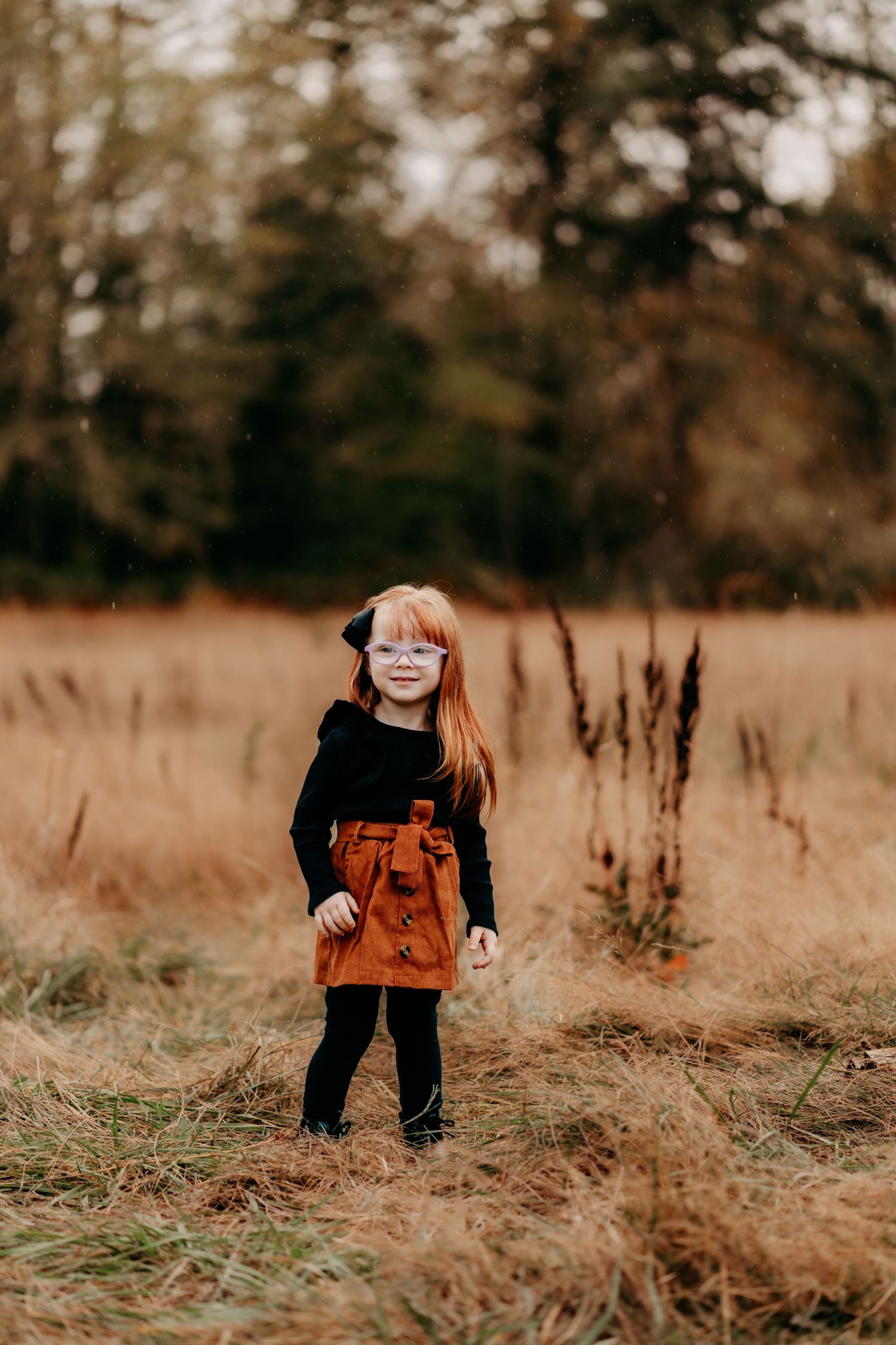 A young red-haired girl in a black top and rust skirt stands smiling in an autumn field during fall photo session in Puyallup, Wa.