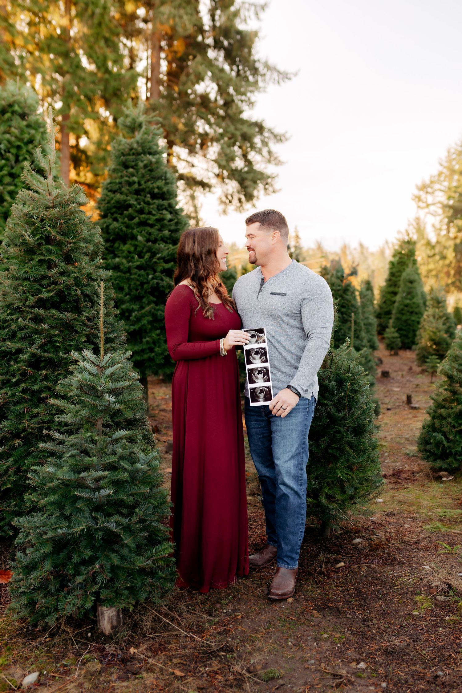 Couple holding ultrasound photos at a Christmas tree farm, woman in red dress, man in gray shirt, surrounded by evergreens.