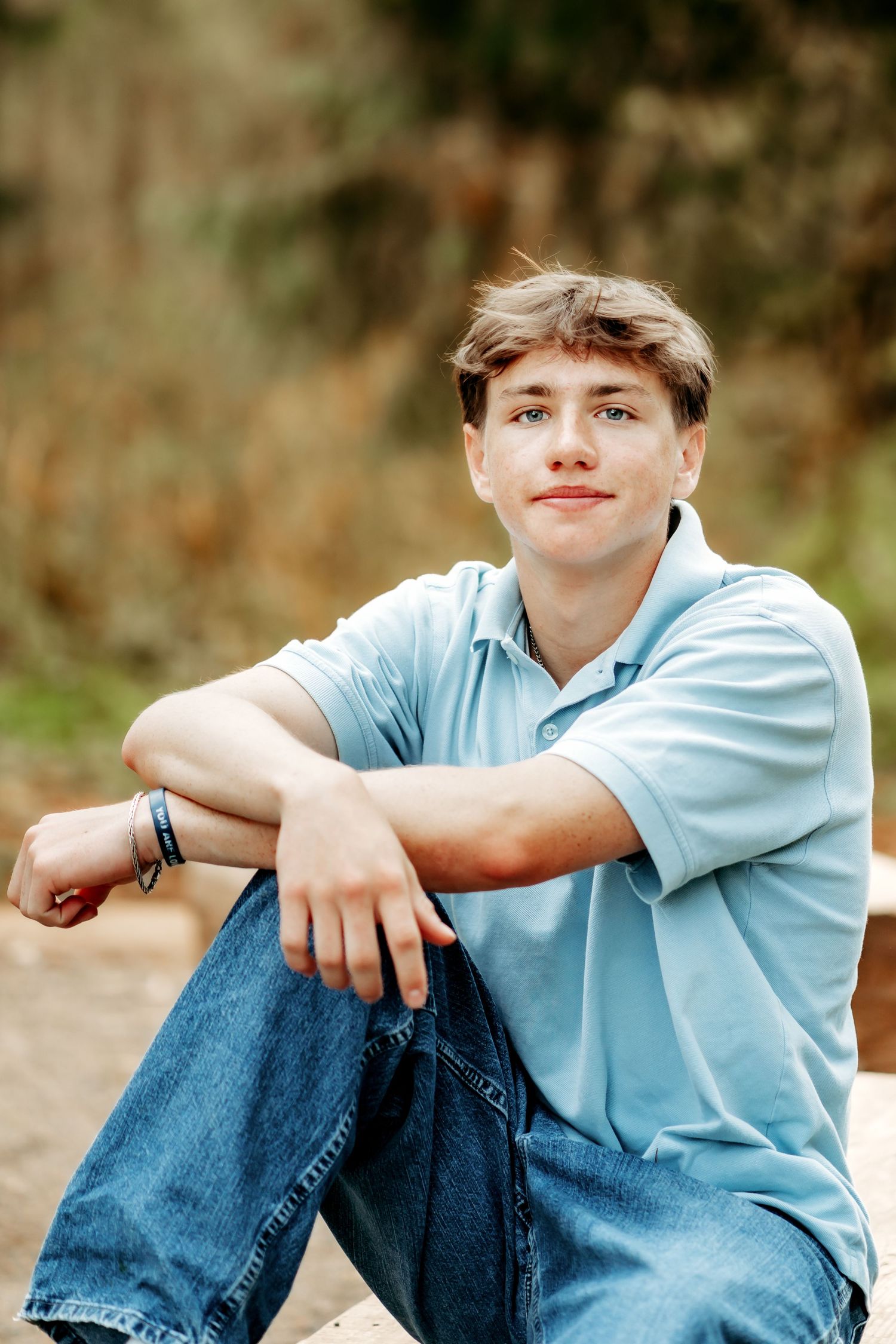 A teenage boy in a light blue polo shirt and jeans sits relaxed outdoors, smiling at the camera during his high school senior photos.