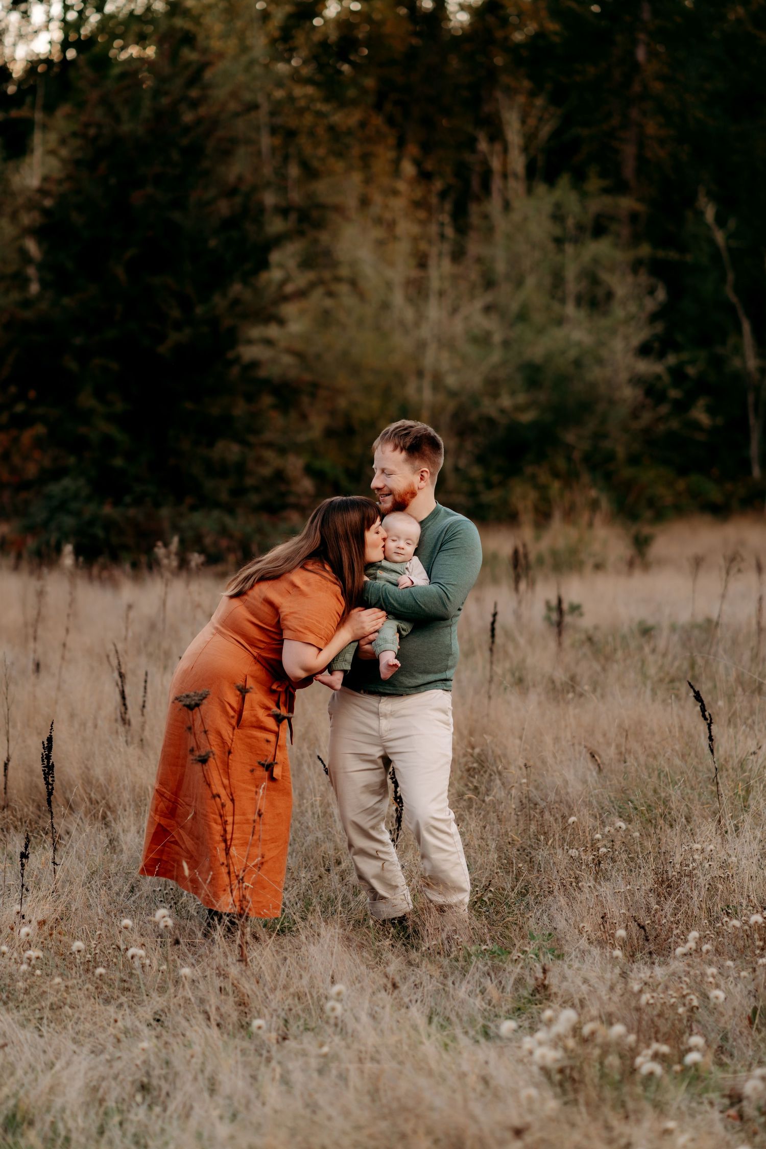 A young family poses in an autumn field, mother in orange dress and father in green sweater holding their baby for family photos in Puyallup
